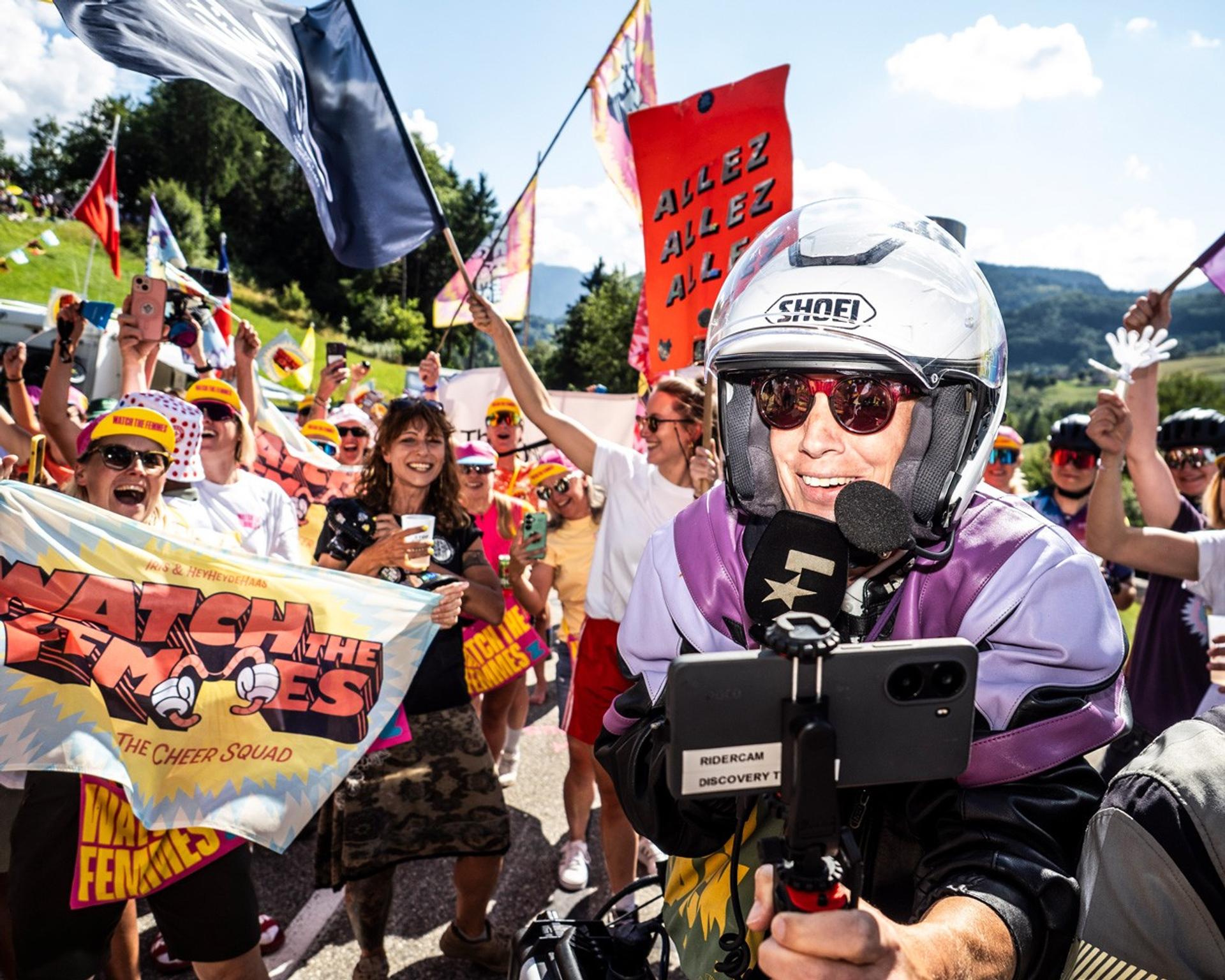 Masked rider in helmet and sunglasses waves to a cheering crowd at a cycling fan event