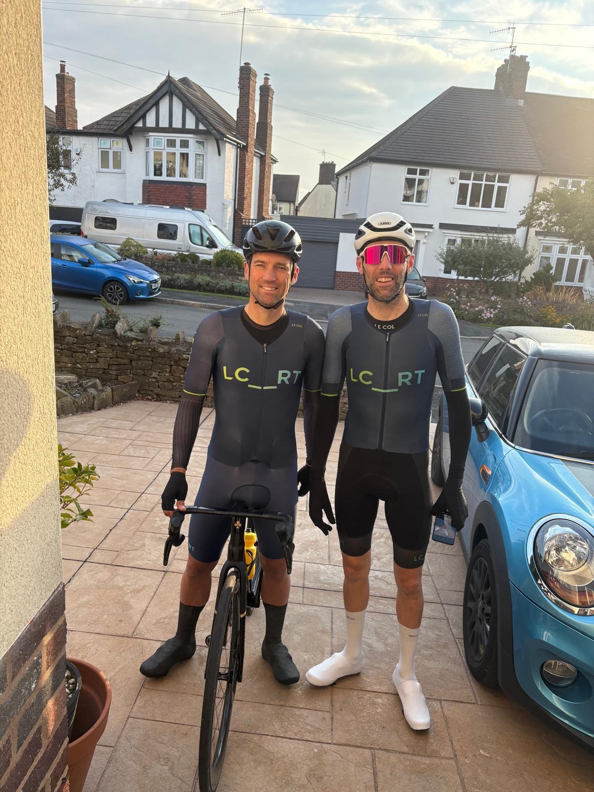 Two cyclists pose with road bikes outside suburban houses, wearing matching black kits and helmets