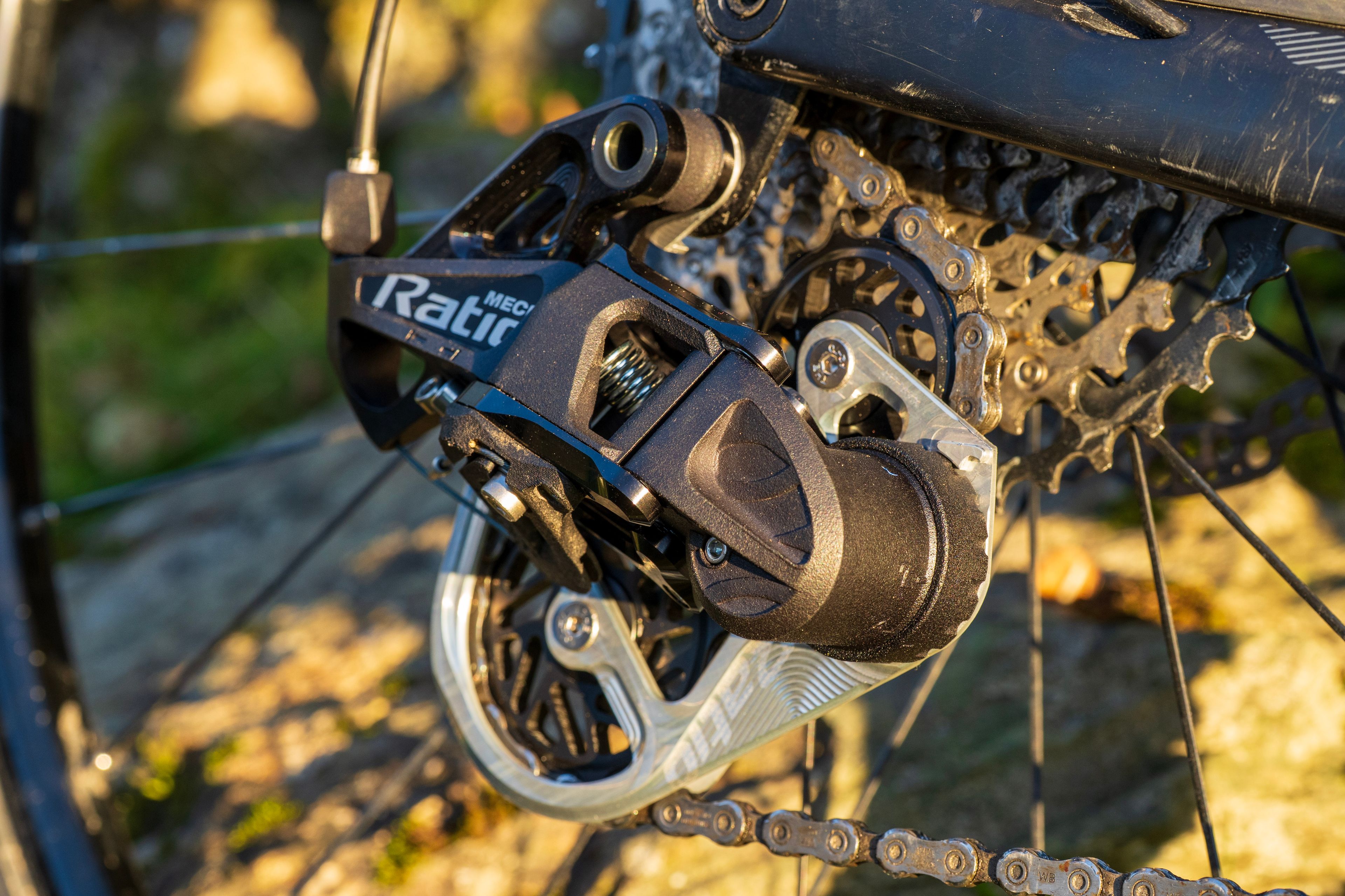 A close-up of a Ratio Mech rear derailleur in use, showing the silver lower cage, clutch housing and jockey wheels, with dirt on the cassette and frame in natural sunlight.