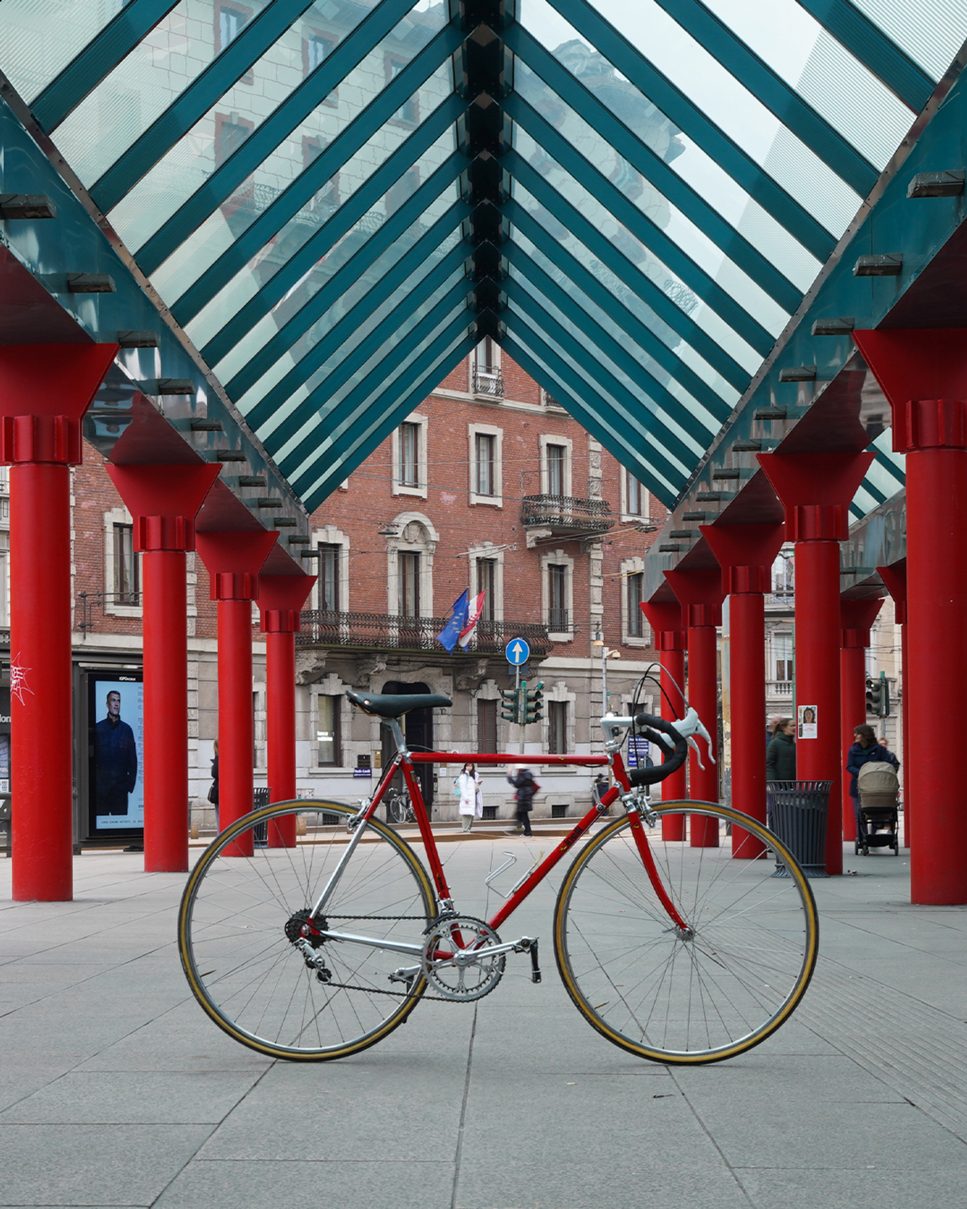 Red Cinelli Supercorsa steel road bike with drop handlebars and rim brakes, photographed under a glass-roofed walkway with red columns in an urban setting.