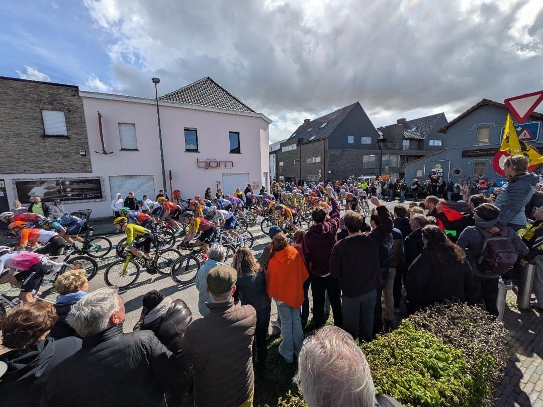 The Tour of Flanders peloton races through a Flemish village as spectators line both sides of the road under dramatic cloudy skies