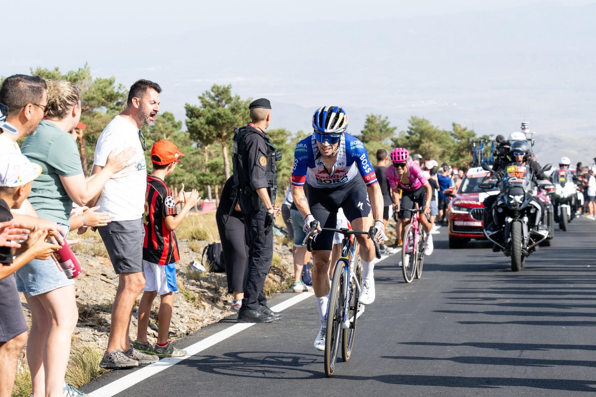 Enric Mas rides along a road as spectators, a race motorcycle and support cars follow behind.