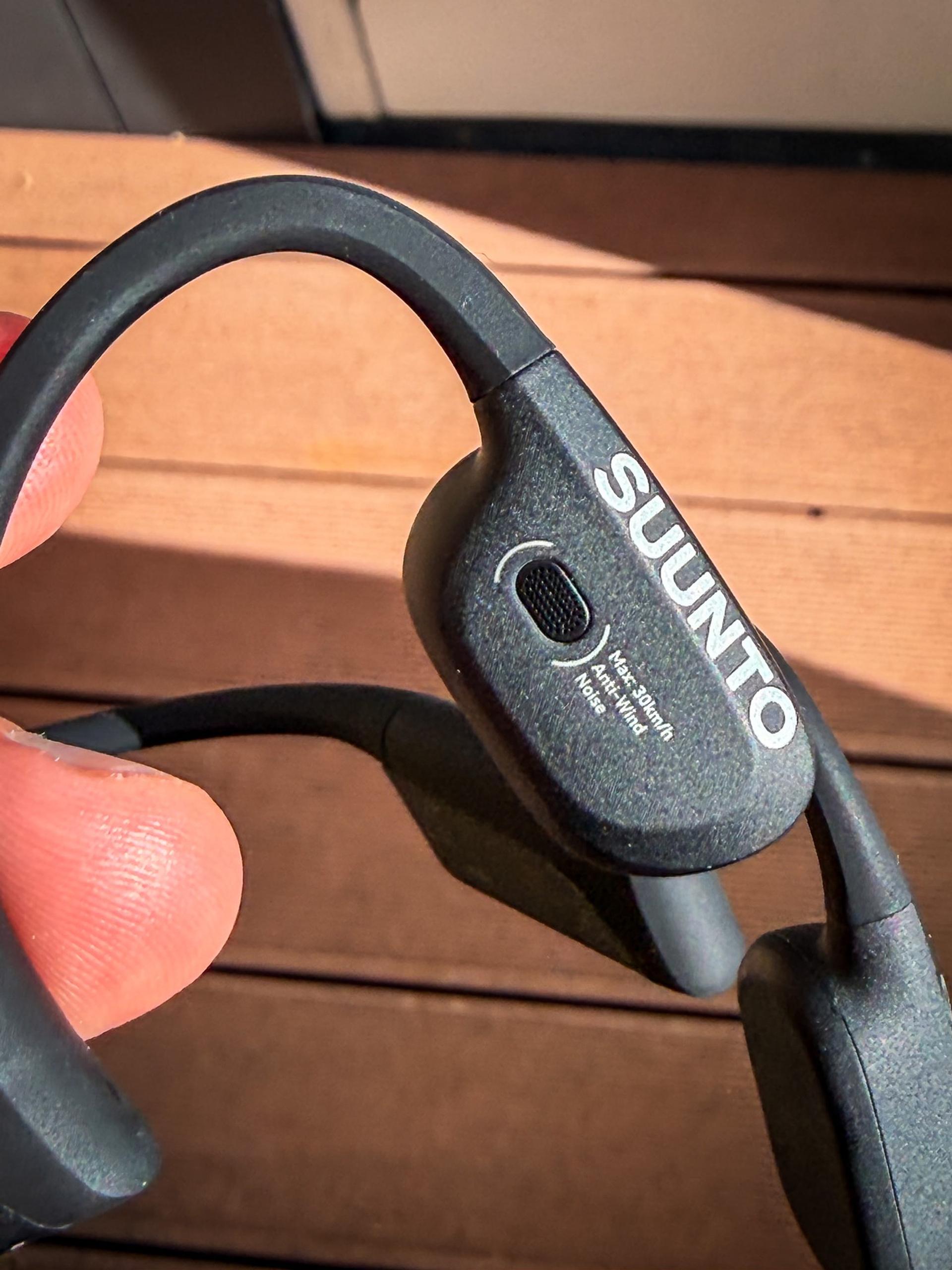A hand holds the dark gray Suunto Wing 2 bone-conduction headset against a wooden tabletop.