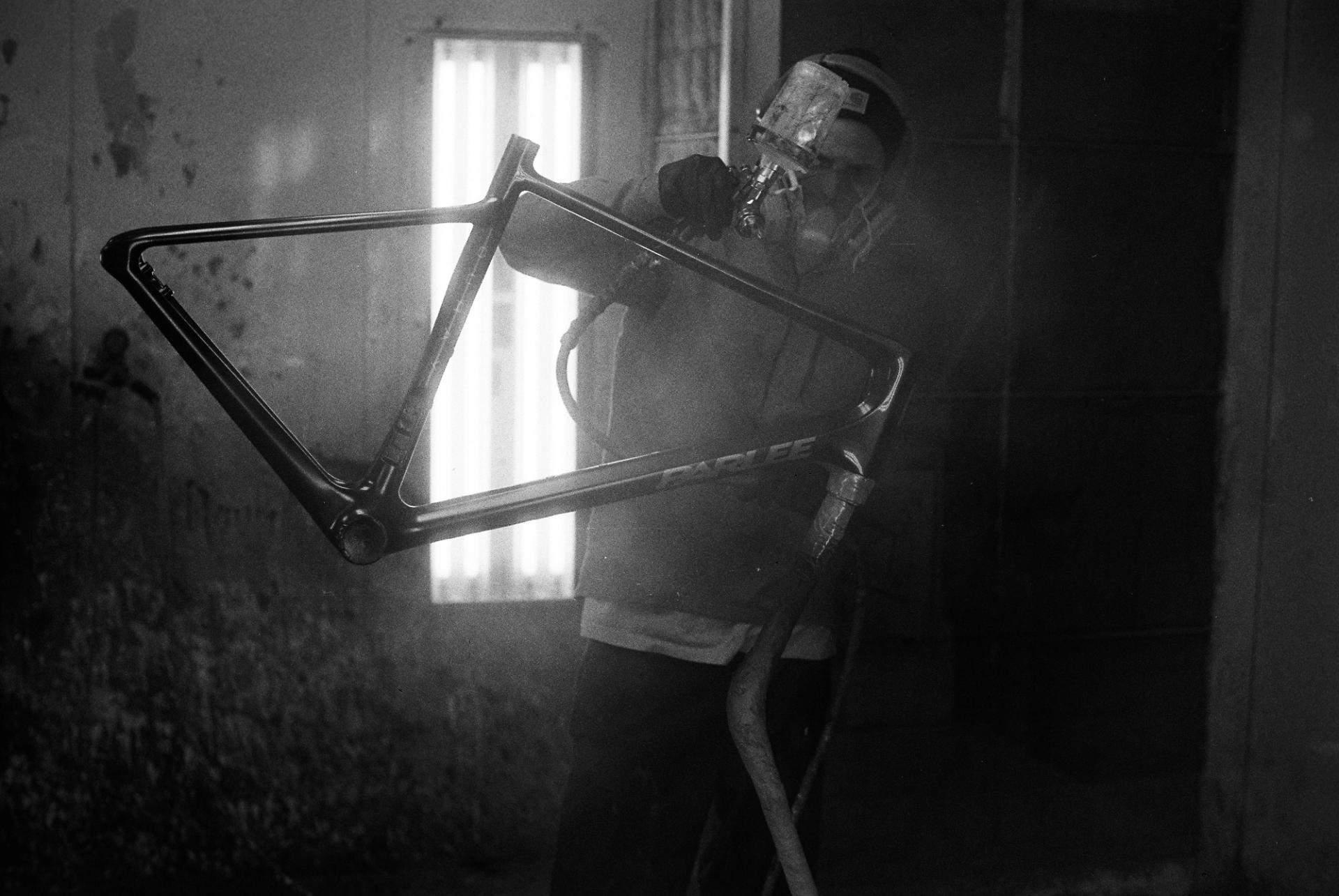 A worker silhouetted against bright window light examines a bicycle frame, the backlit scene lending a contemplative quality to the moment.