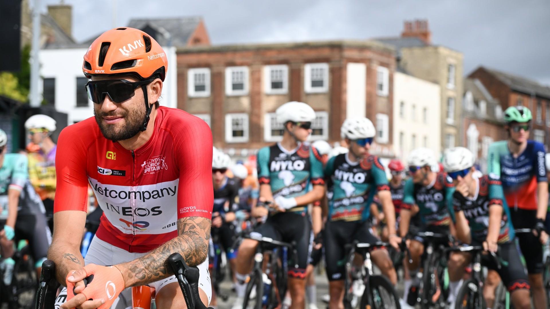 Filippo Ganna in a red Netcompany INEOS jersey rides at the front of a group of cyclists on a city street.