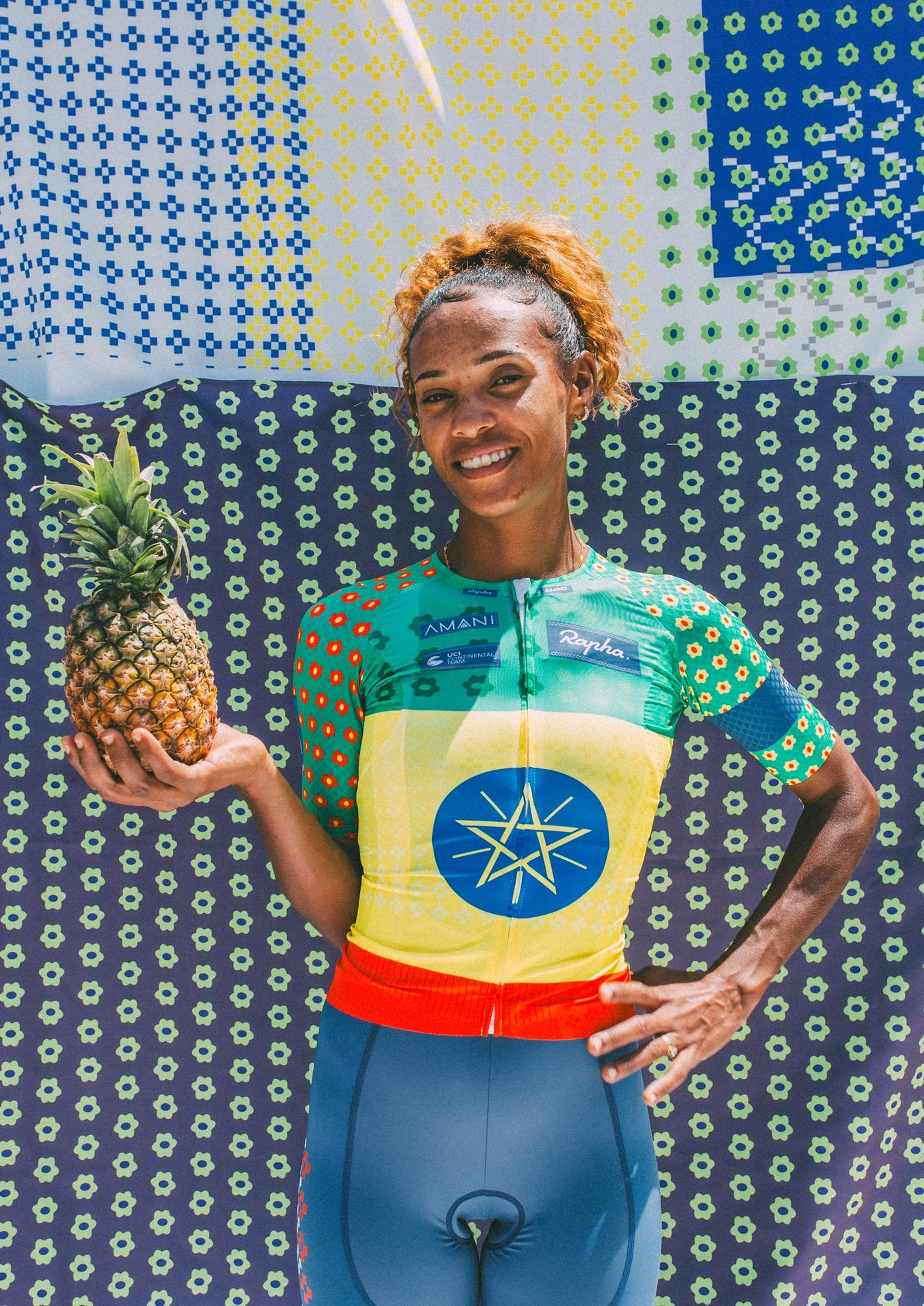 Cyclist in colorful jersey celebrates while holding a pineapple at a tropical-themed podium backdrop