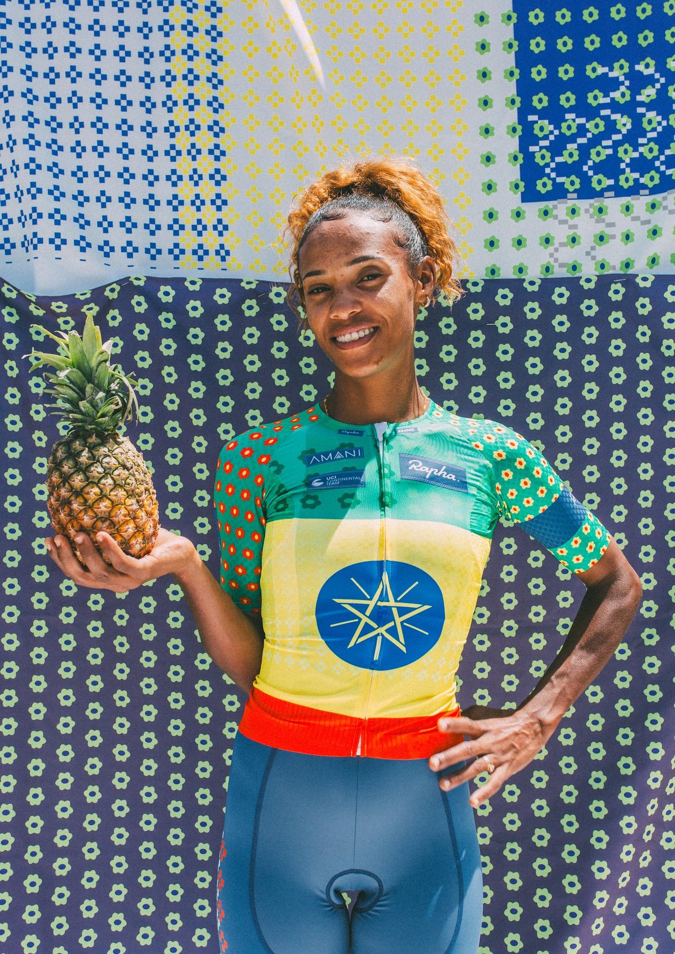 Cyclist in colorful jersey celebrates while holding a pineapple at a tropical-themed podium backdrop