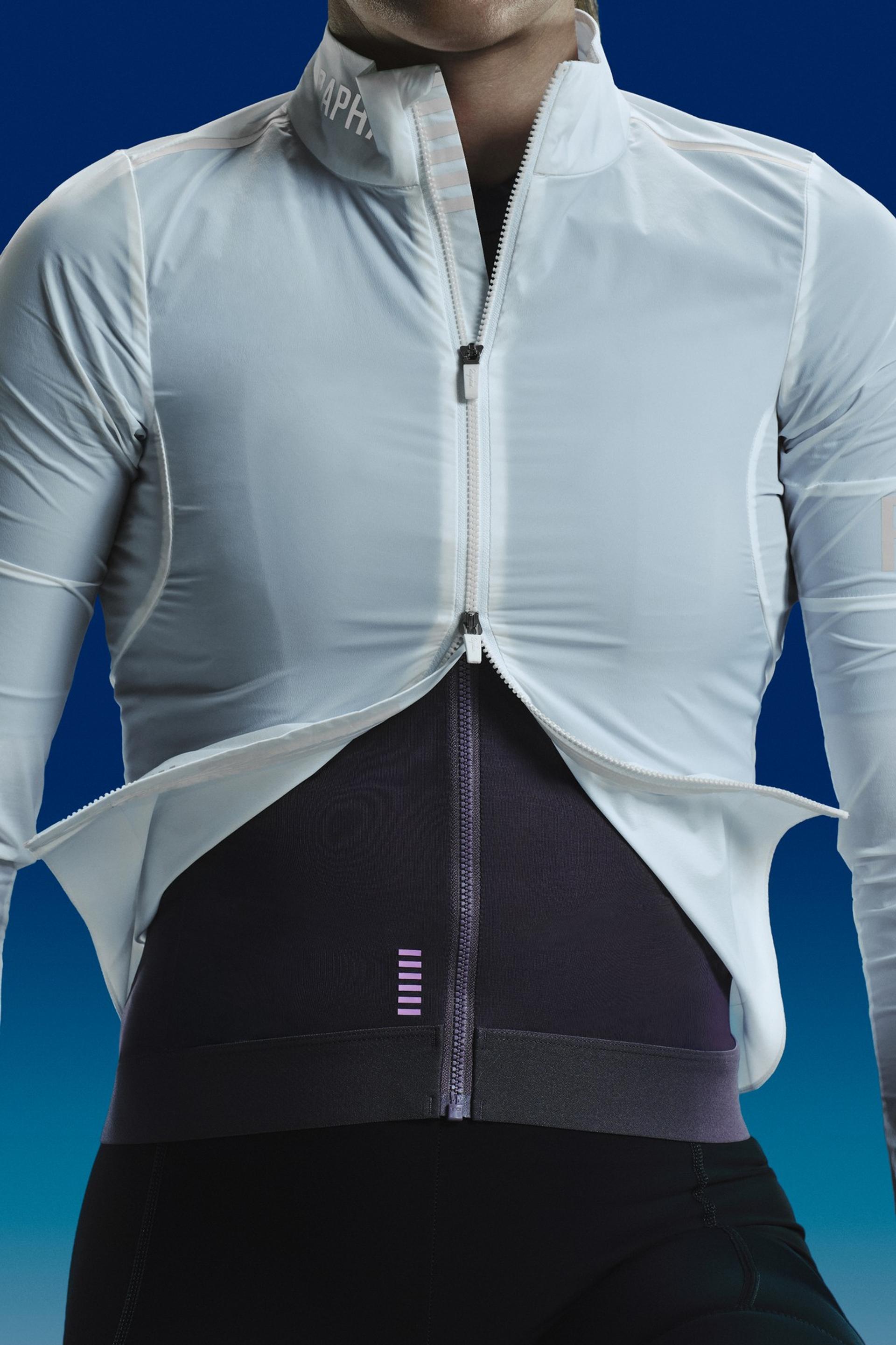 A cyclist wearing a white Rapha lightweight jacket over a purple jersey, partially unzipped from the bottom.
