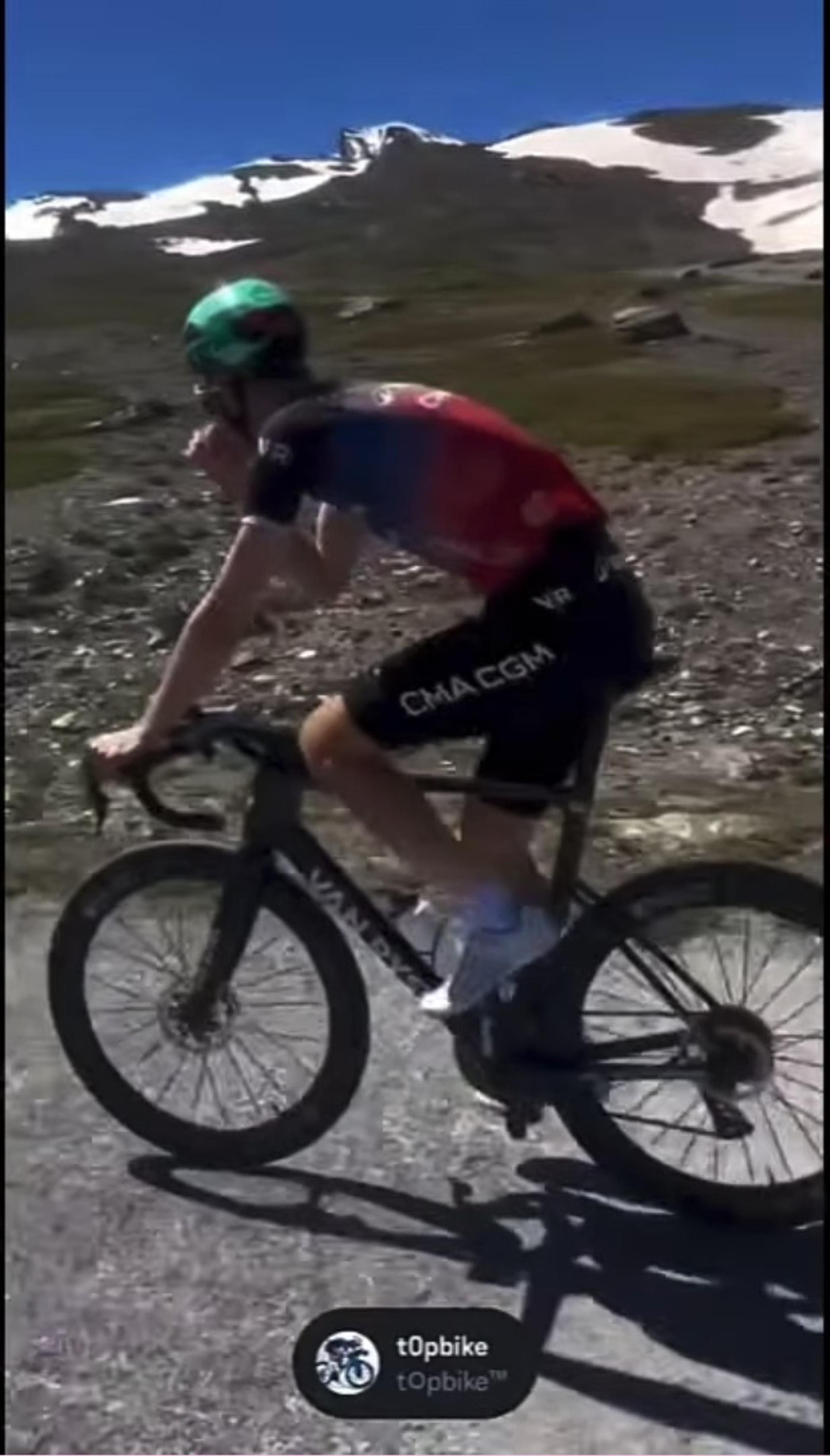 Cyclist in red-and-blue kit climbs a mountain road on a gravel bike under bright sun