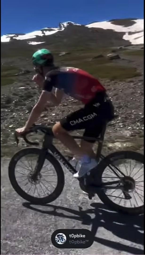 Cyclist in red-and-blue kit climbs a mountain road on a gravel bike under bright sun
