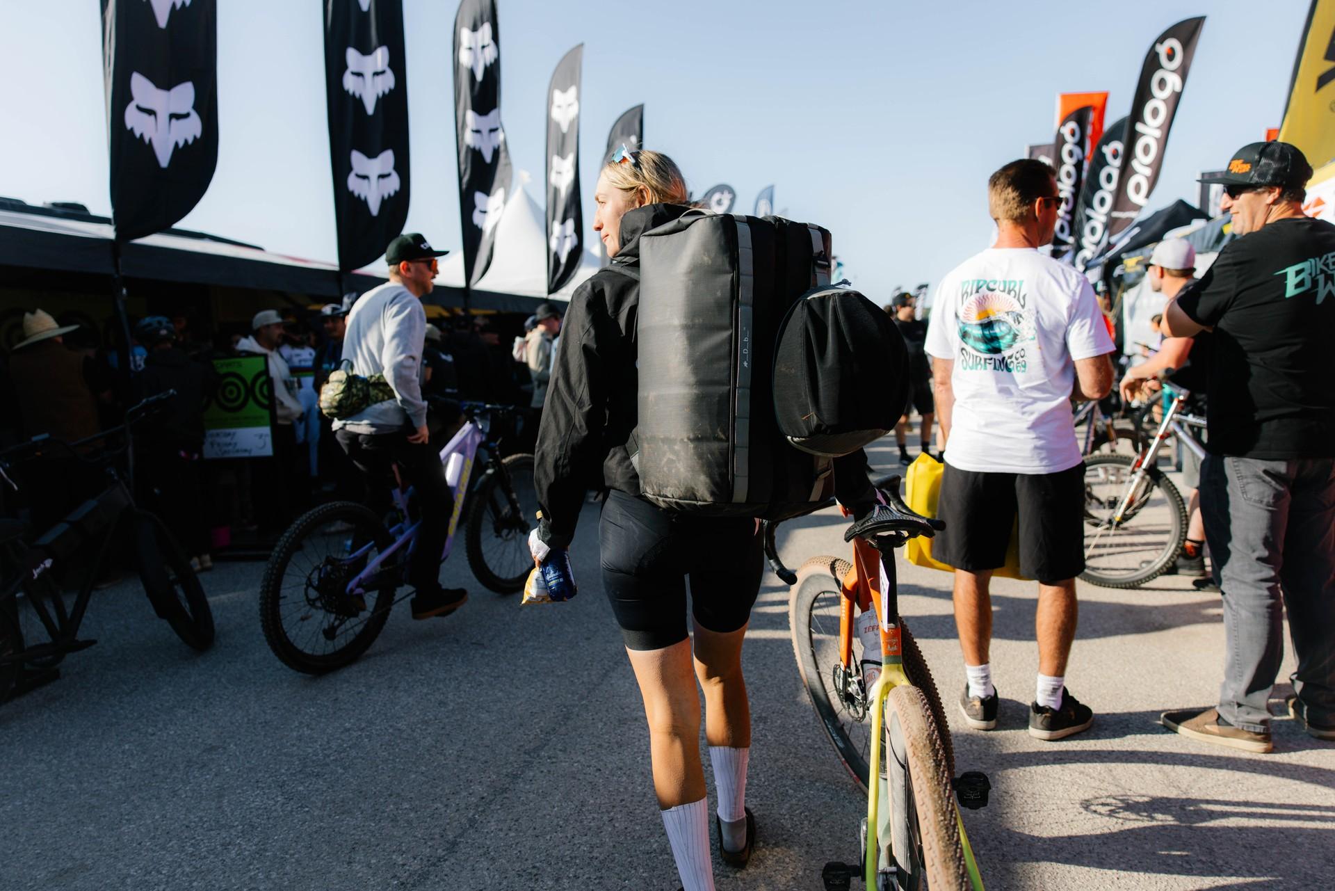 Cyclists gather beside bikes and sponsor banners at a road race event in bright afternoon light.