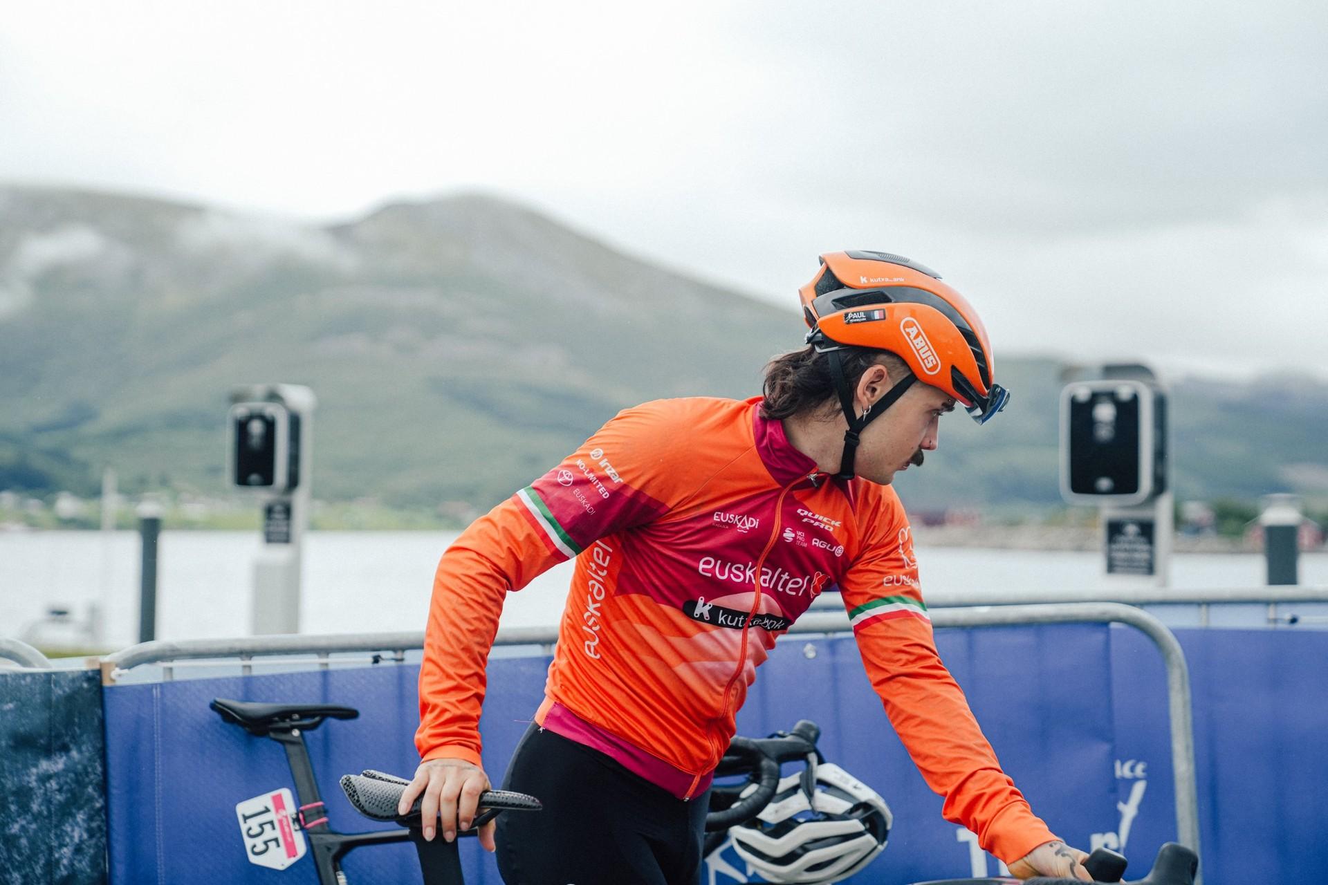 Euskaltel-Euskadi cyclist in an orange team jersey and helmet leans over a bike beside a blue barrier.