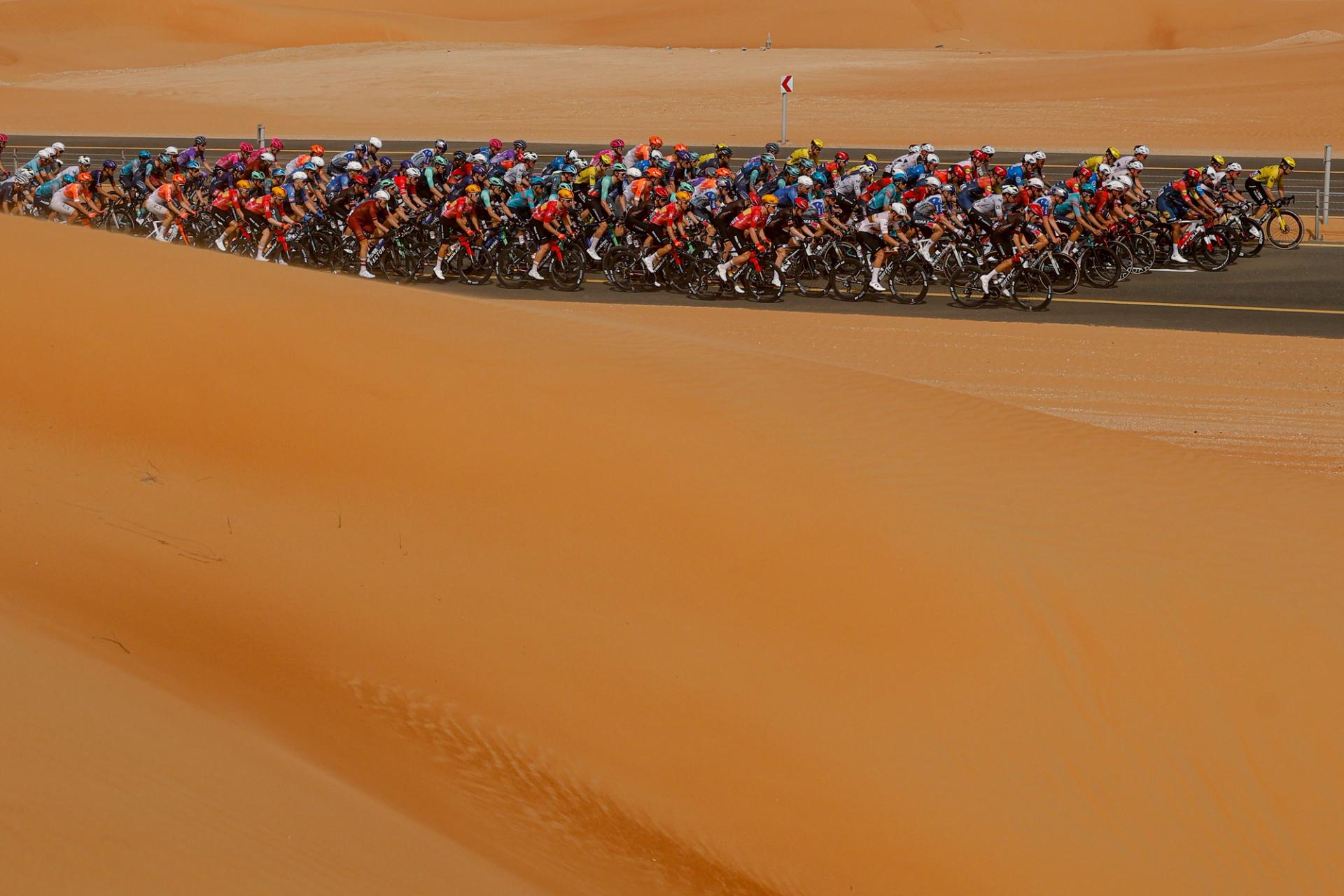 Peloton of professional road cyclists racing along a paved desert road, tightly packed as they pass between large orange sand dunes under clear skies.