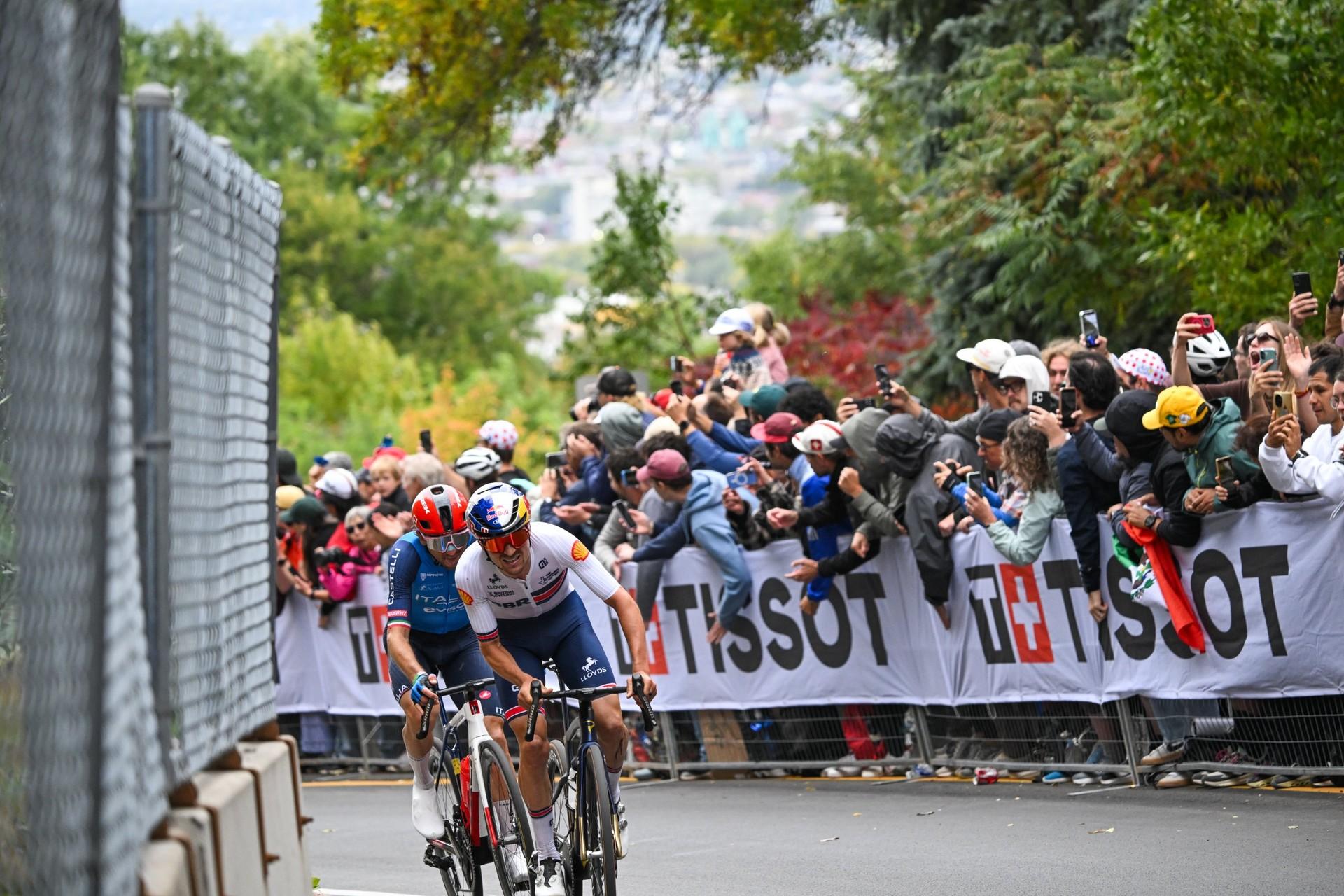 Road cyclists race past Tissot-branded barriers, with spectators watching behind them.