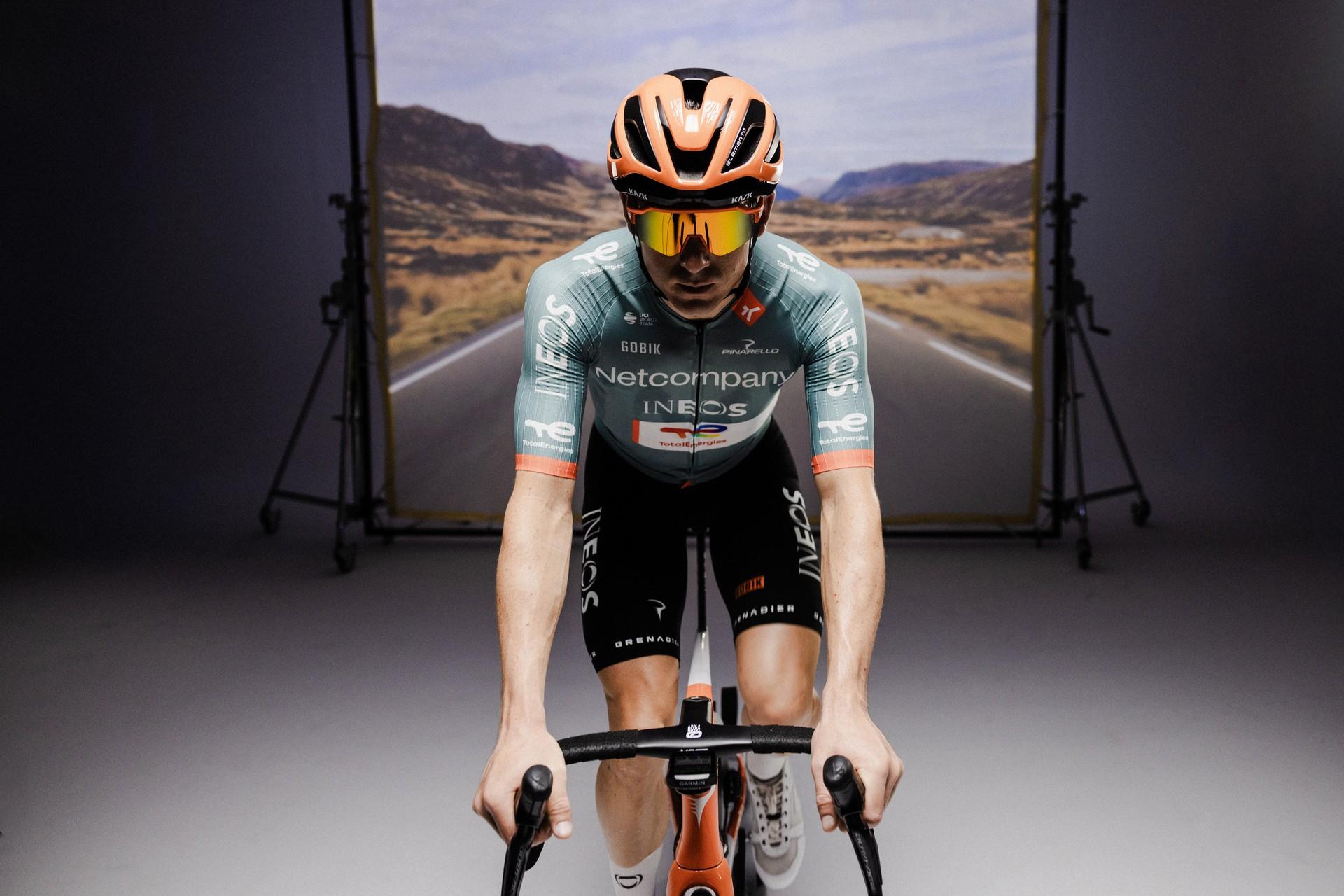 Cyclist in INEOS Grenadiers kit rides forward on a studio stage with mountains in the background