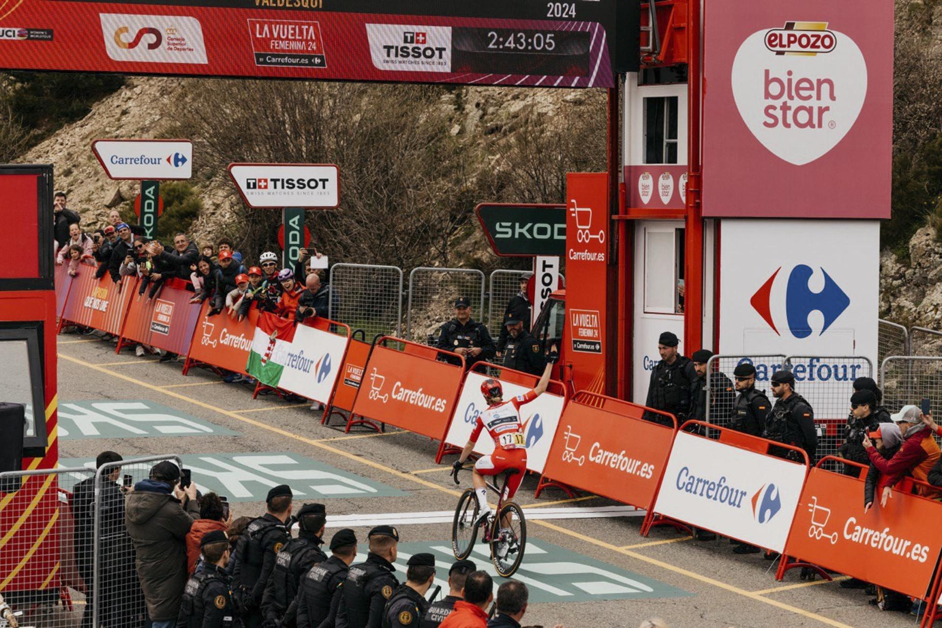 A cyclist in a red and white kit raises an arm as she crosses the La Vuelta Femenina finish line, with crowds behind the barriers