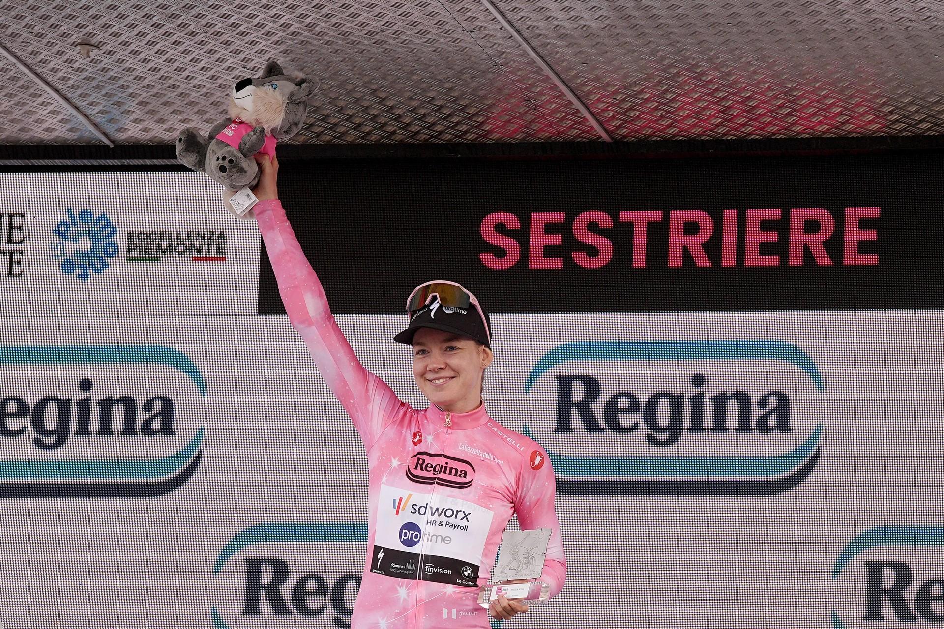 Cyclist celebrates on the podium at an Italian race finish, raising one arm in a victory gesture.