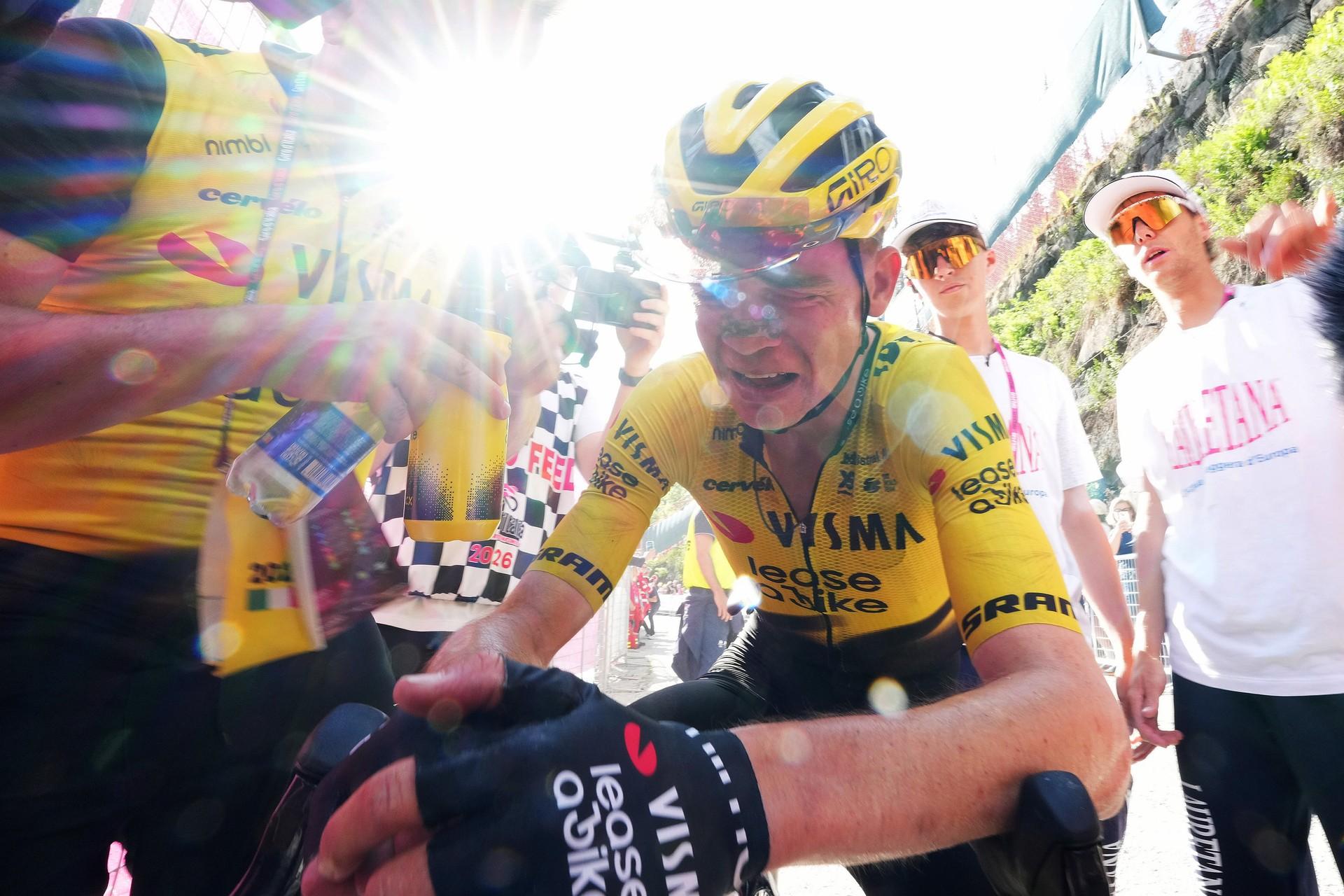 Cyclist in yellow VIVA-VESSEL face mask raises trophy amid cheering team after roadside race