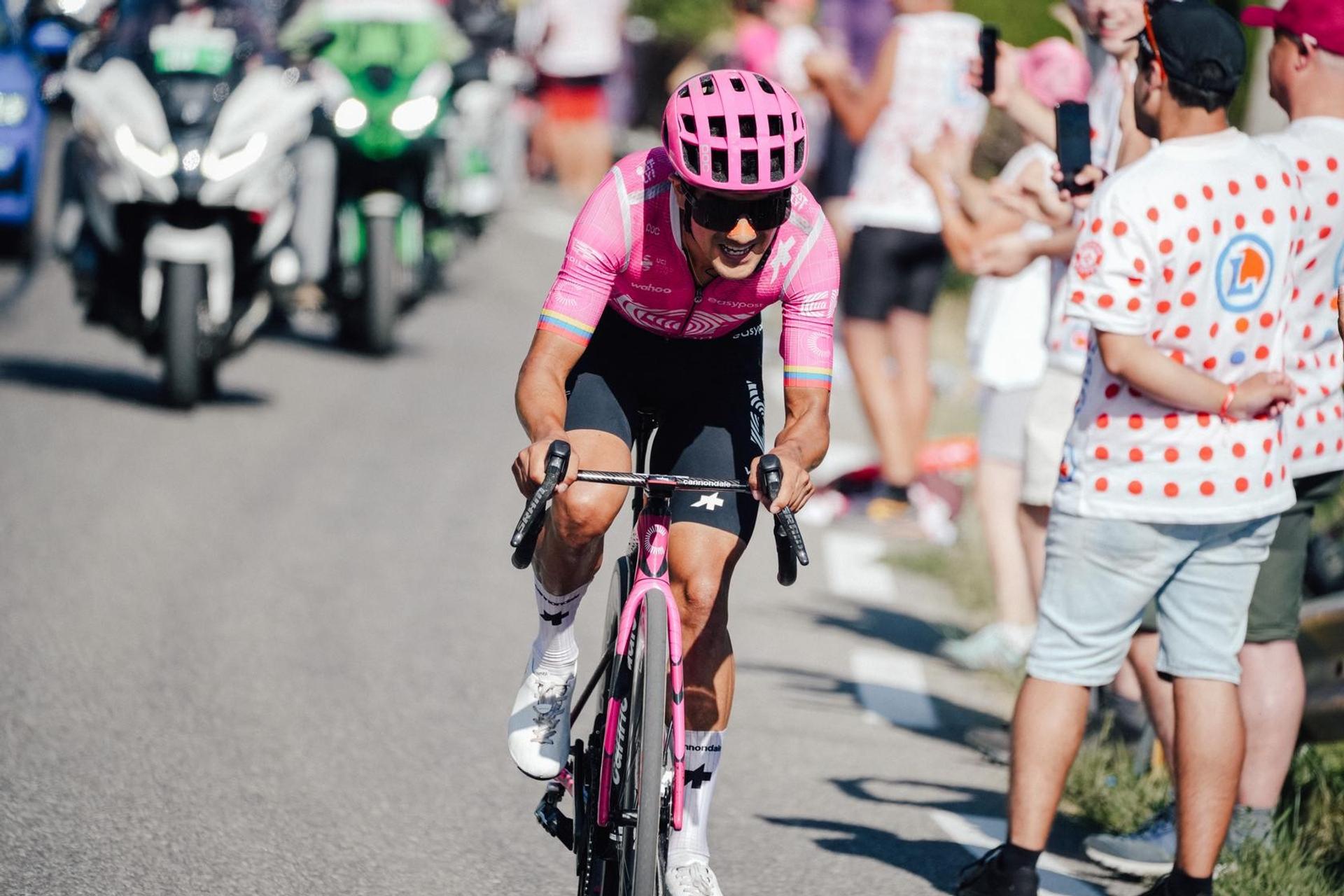 Cyclist in a pink jersey climbs a road past cheering spectators and race motorcycles.