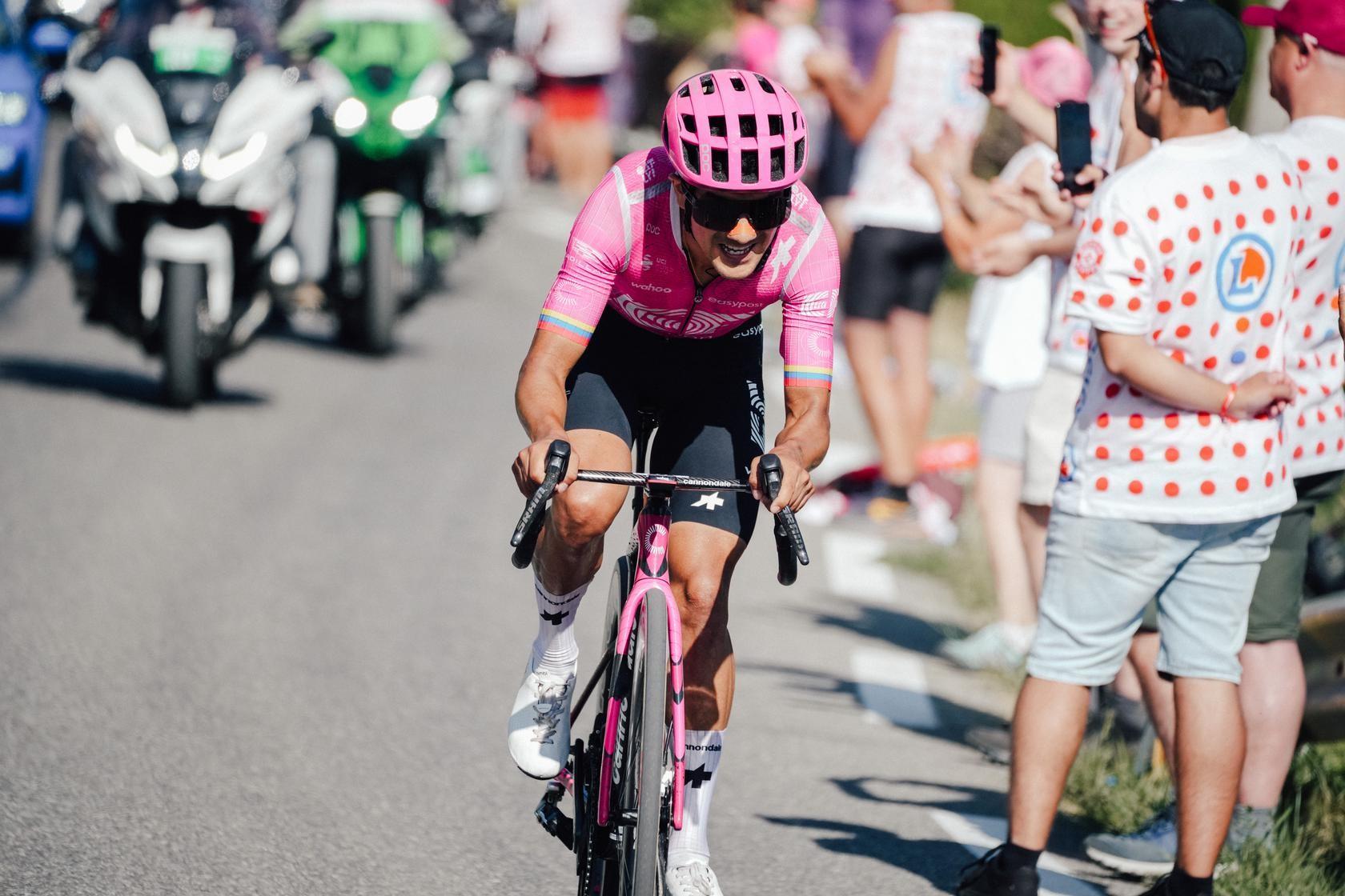 Cyclist in a pink jersey climbs a road past cheering spectators and race motorcycles.