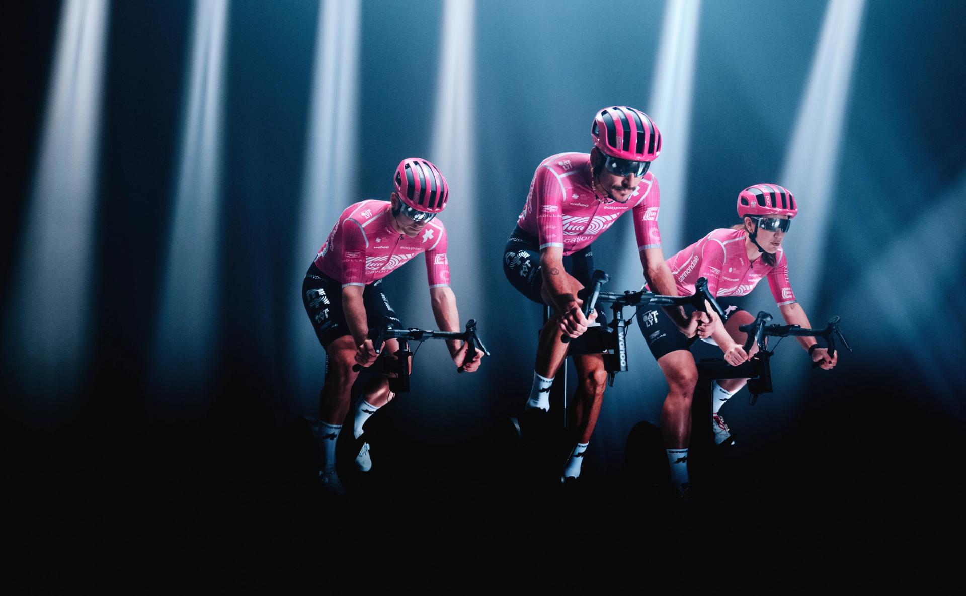 A group of professional cyclists in matching pink team kits and helmets riding stationary bikes under dramatic spotlights, with a dark background fading into shadow.
