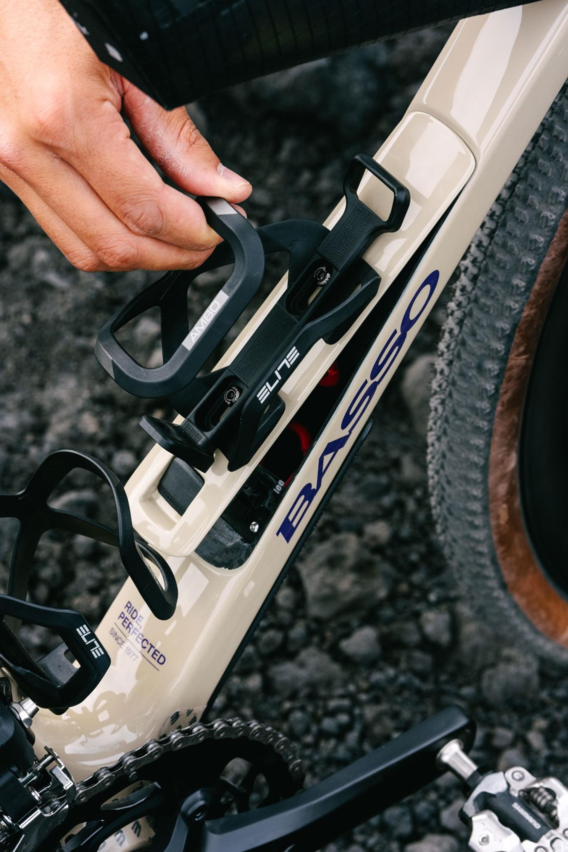 A hand adjusting a black Elite bottle cage on a beige Basso gravel bike frame.