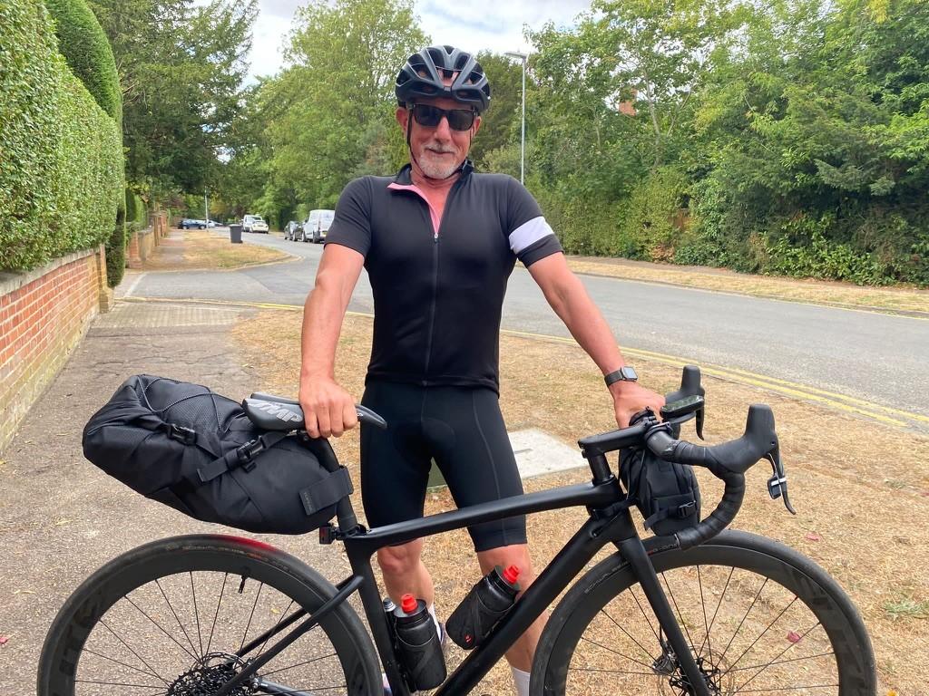 Ewan Kirk in cycling kit stands behind his loaded gravel bike on a quiet road, wearing a helmet and sunglasses.
