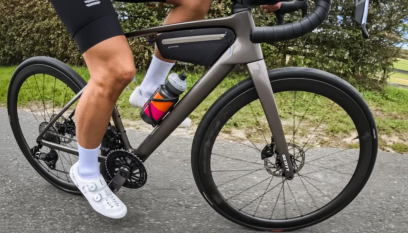 A cyclist rides a Scott road bike with a frame bag and white Shimano shoes along a paved road.