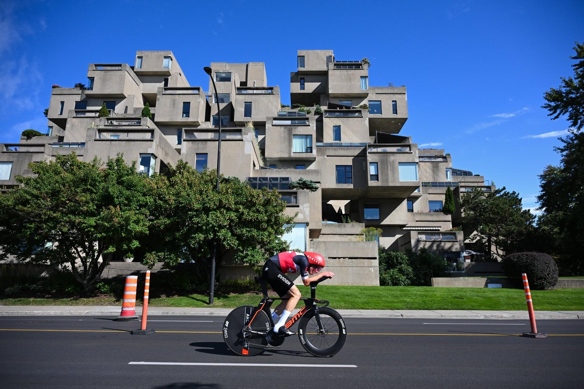 A cyclist in a black-and-red skinsuit rides a time trial bike past cones on a city street.