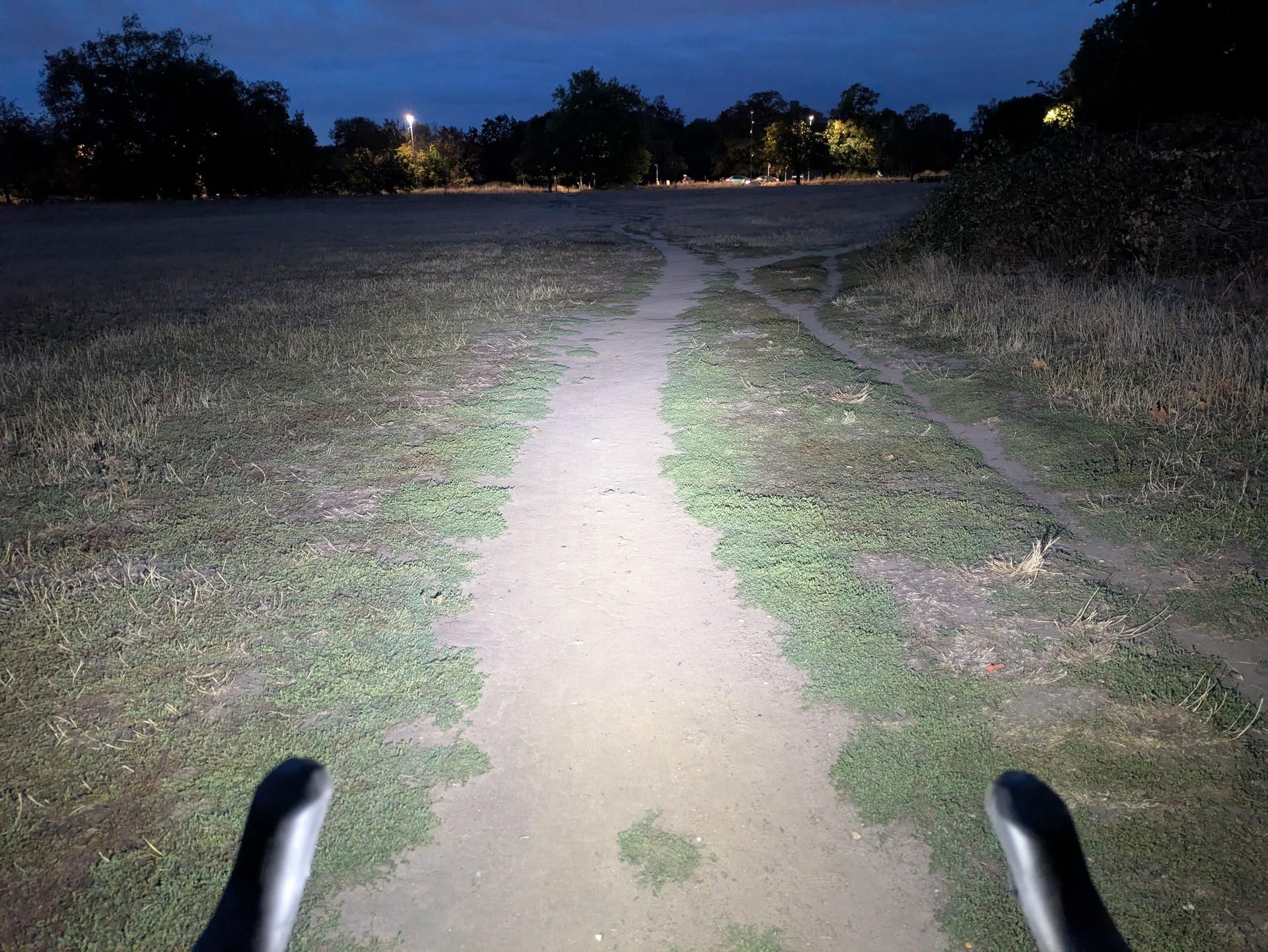 A Exposure Strada RS bike light illuminates a dirt track at night, with handlebars visible in the foreground and trees beyond.