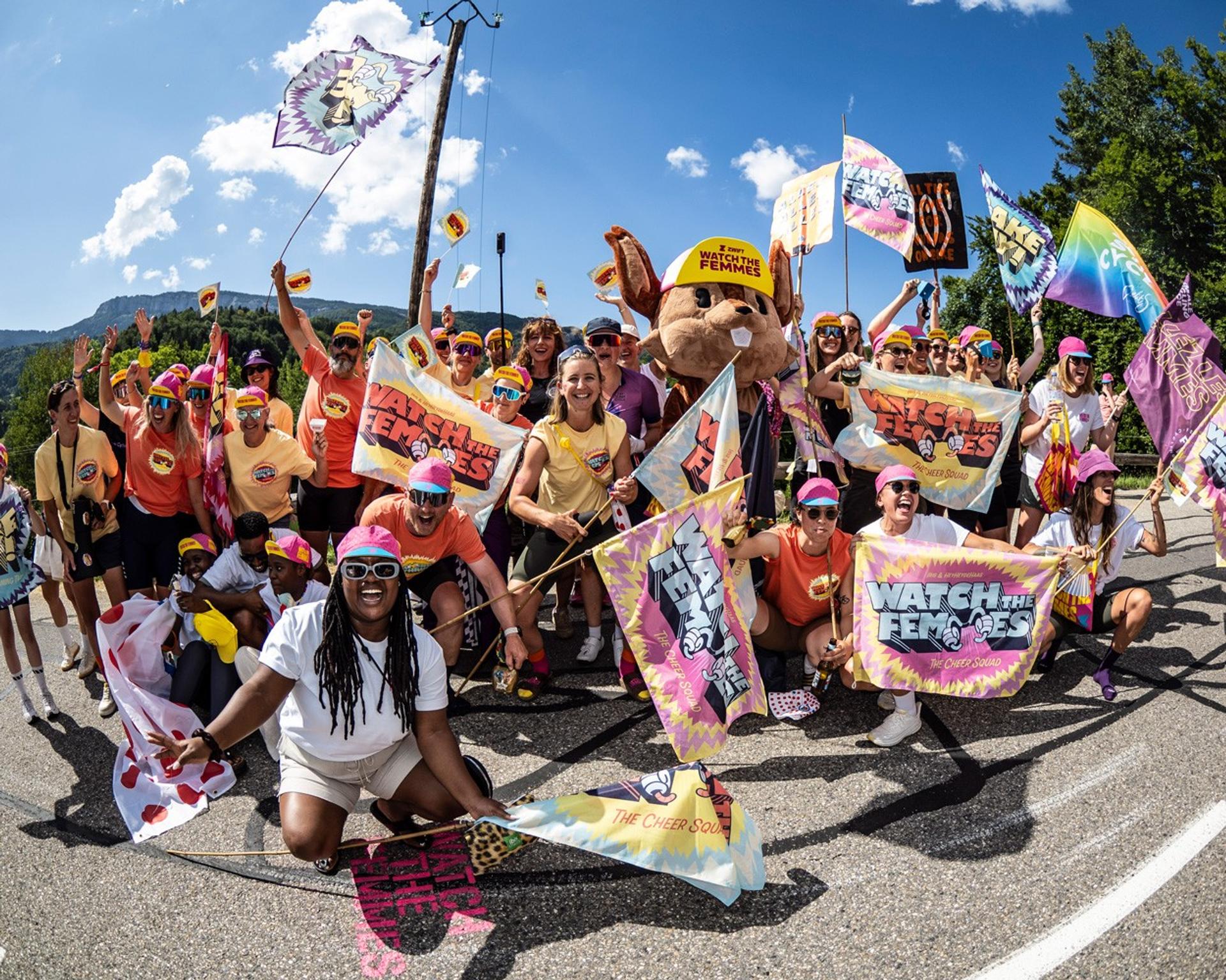 Cycling fans and riders pose together with colorful banners on a sunny road.