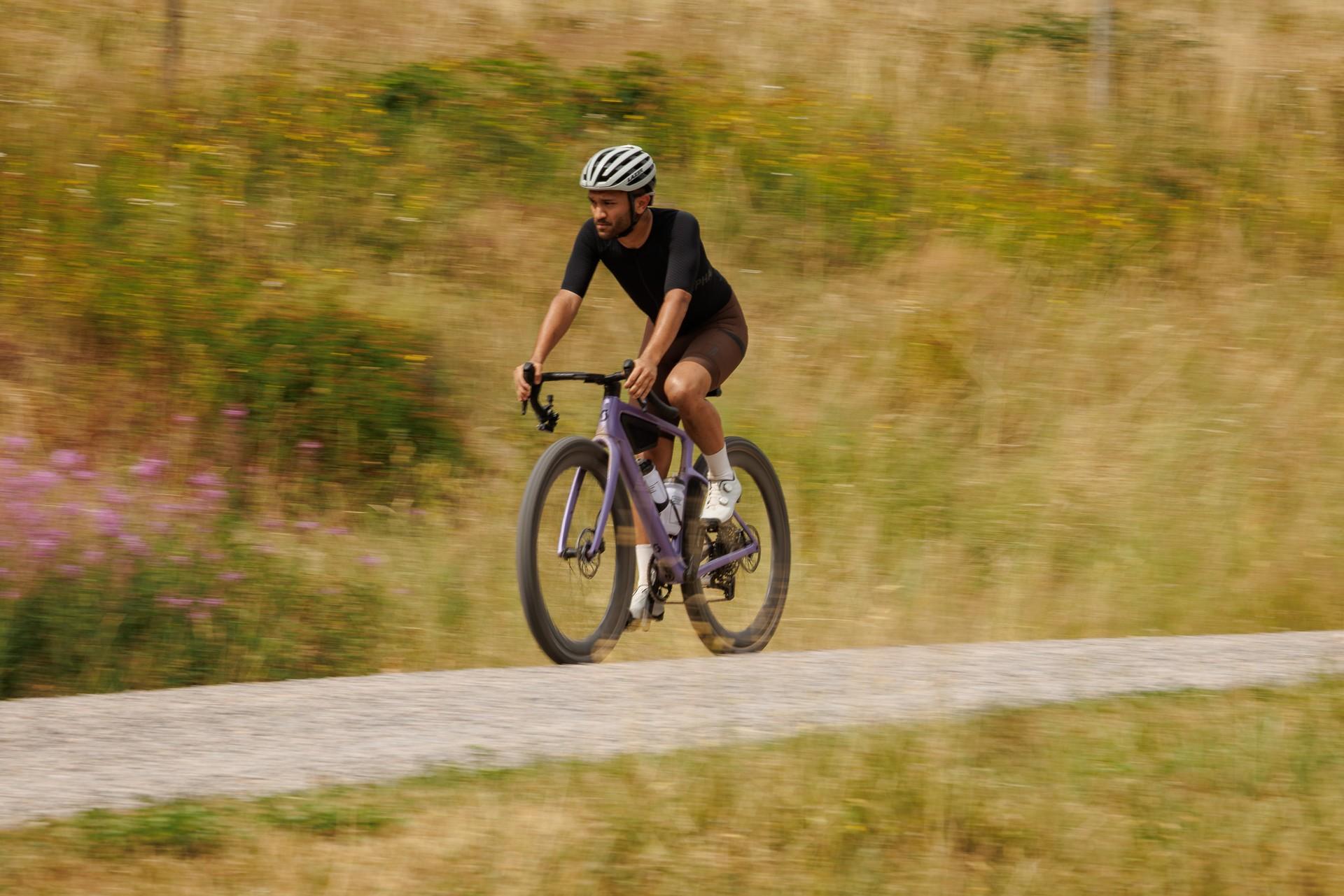 Cyclist in a helmet rides a gravel bike along a gravel path through a grassy meadow.