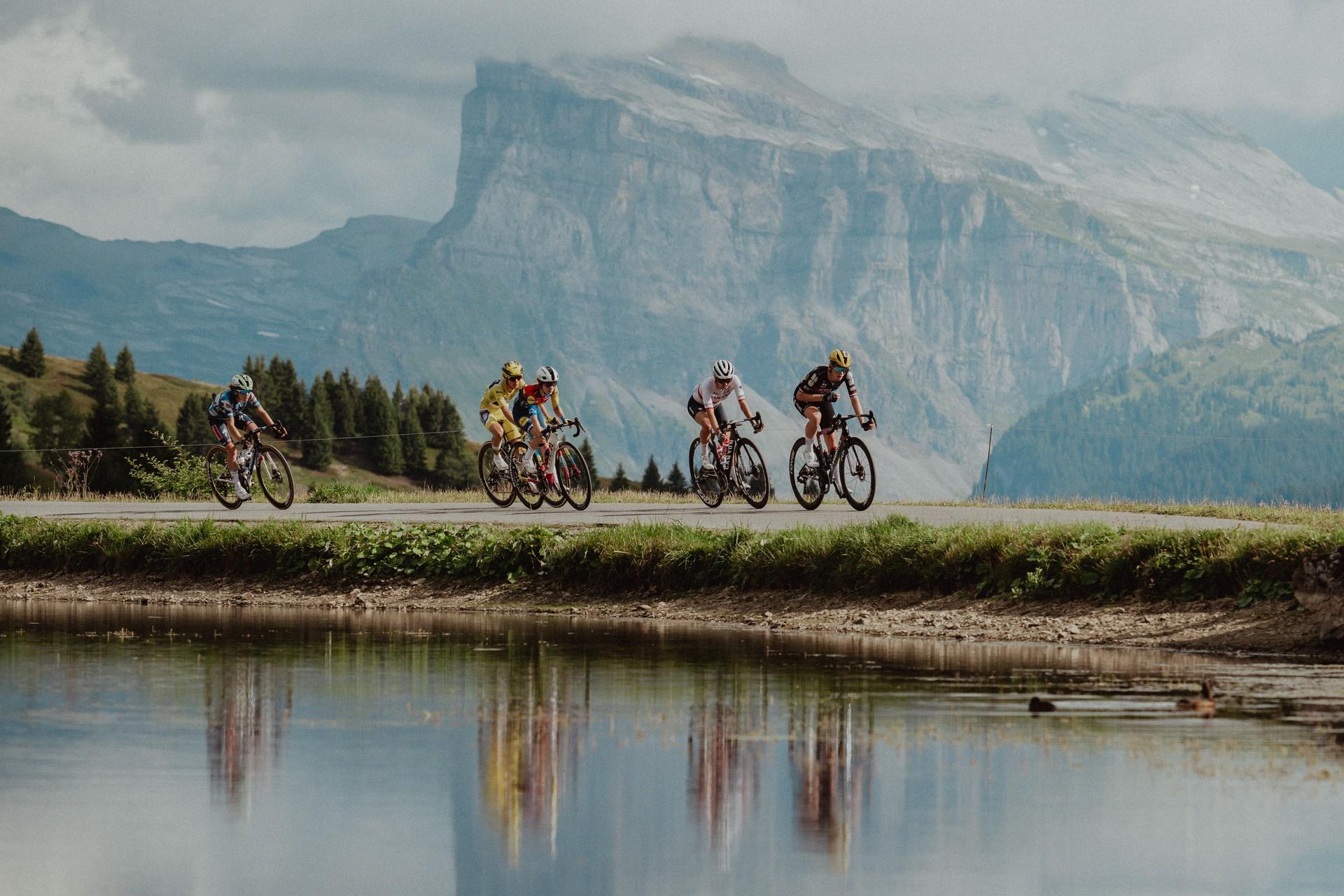 Cycling peloton rides along a mountain lake during Tour de France Femmes avec Zwift stage 9