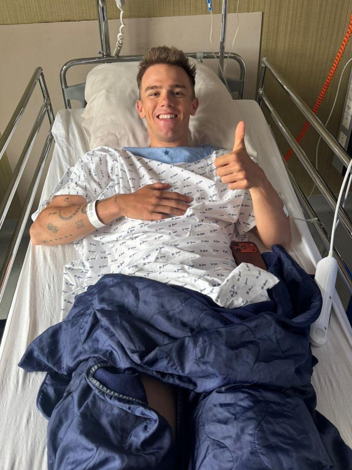 Thibau Nys smiling and giving a thumbs up while lying in a hospital bed wearing a medical gown.