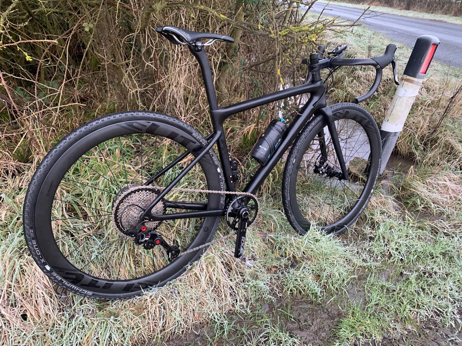 Ewan Kirk’s black gravel bike with drop handlebars and disc brakes standing in grass beside a hedge.