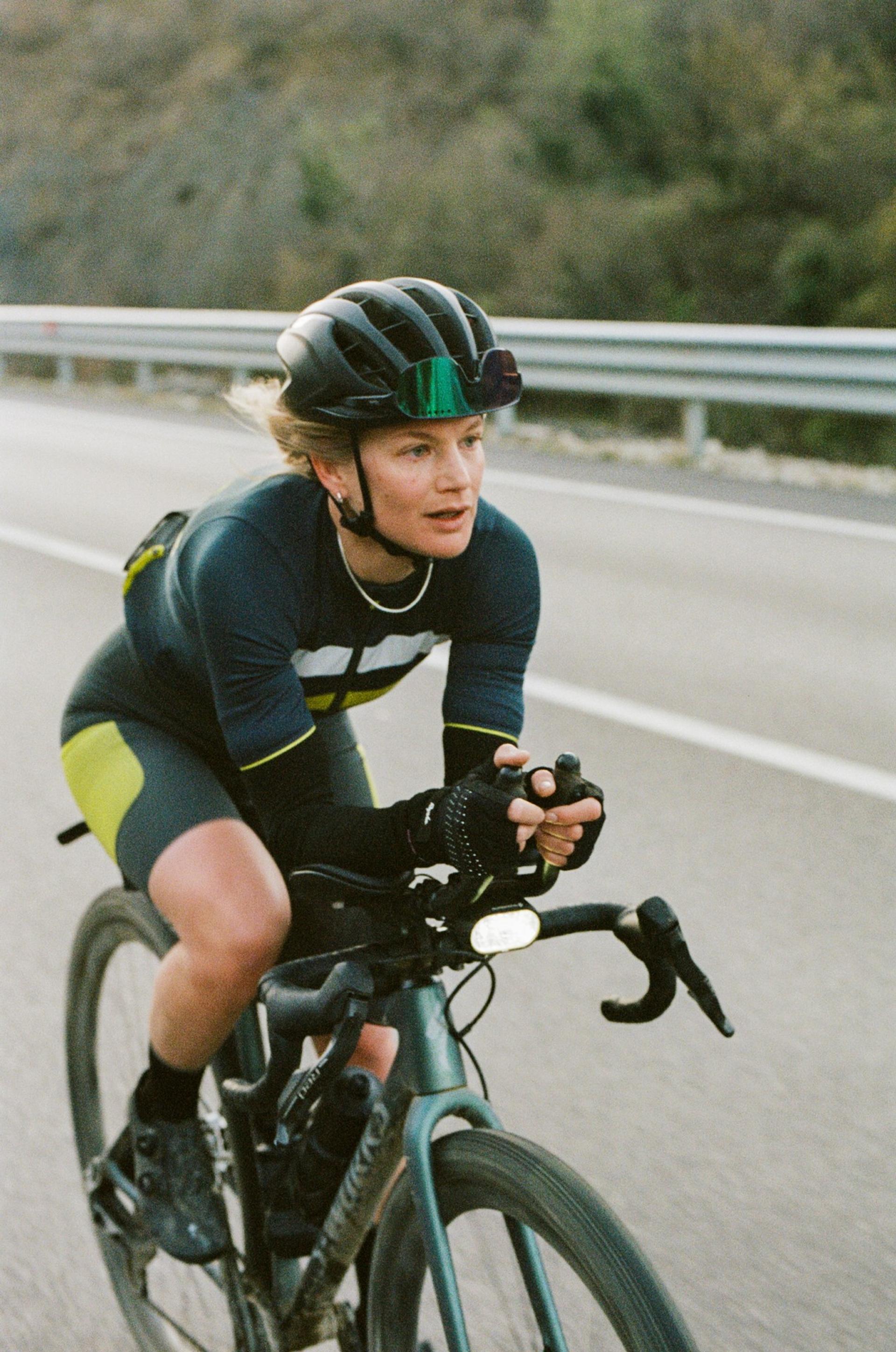 Cyclist in teal-and-black kit rides a road bike while gripping the bars during a race on a hillside road