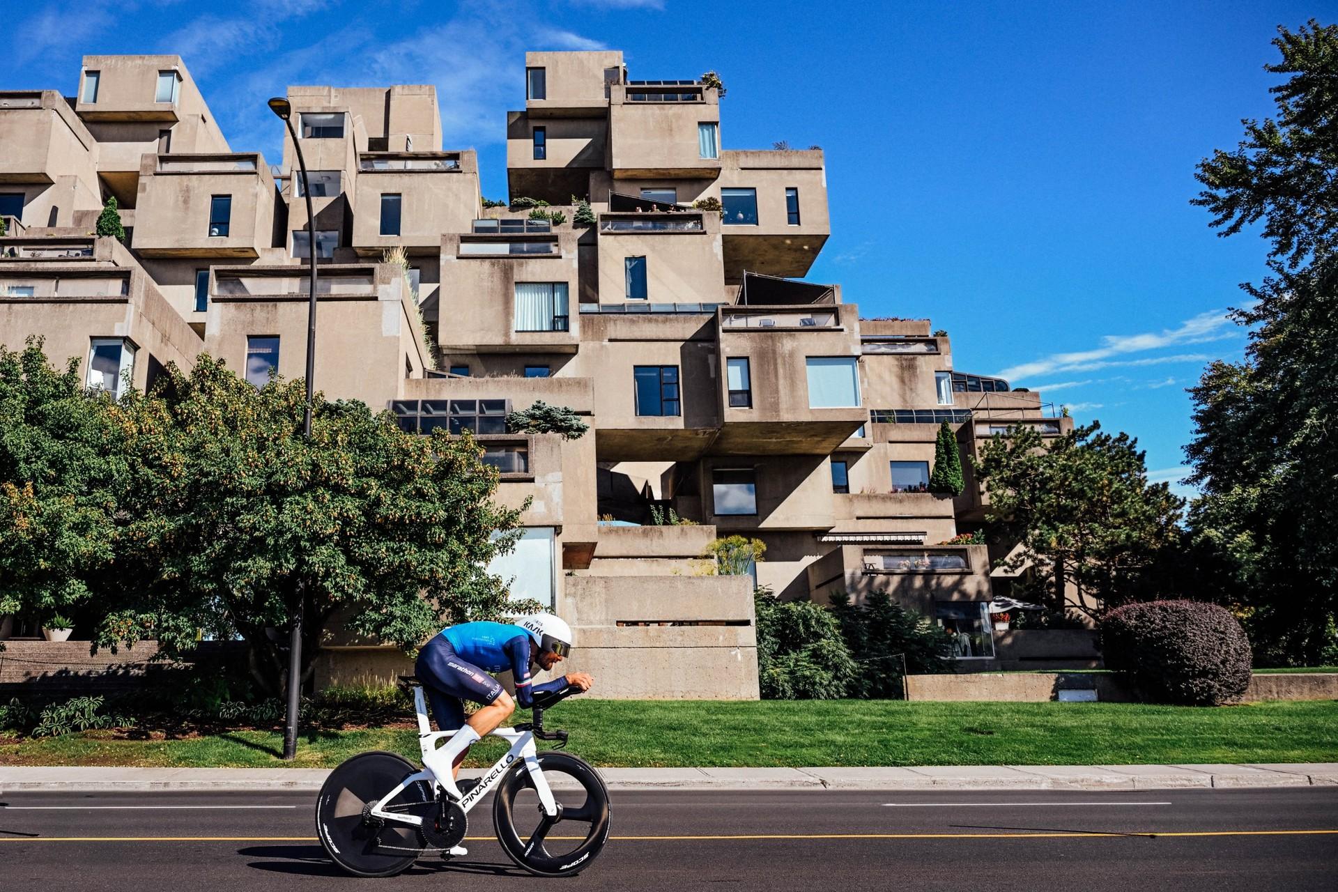 Cyclist in a blue jersey rides a time trial bike along a city street past apartment buildings.