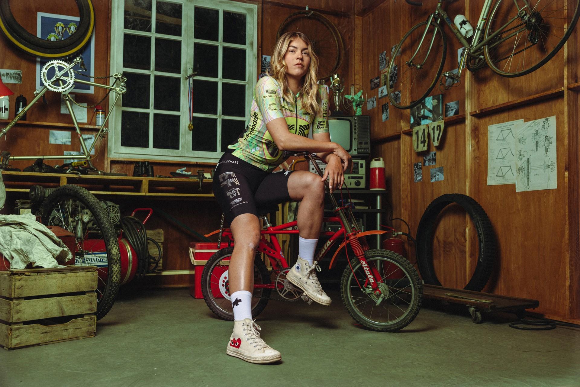 Cyclist in indoor workshop sitting on a red mountain bike, posing near training tools and banners.