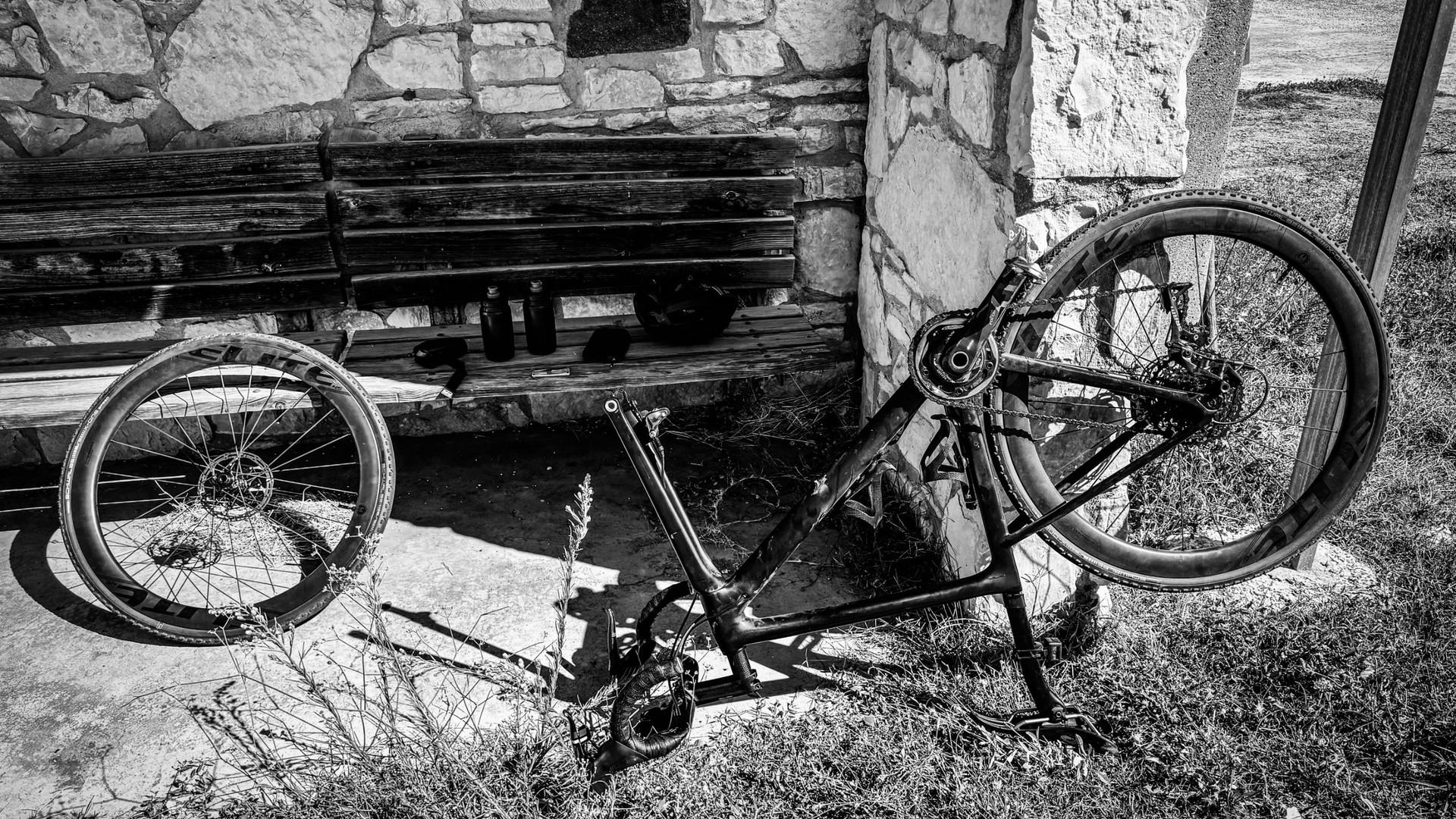 A black gravel bike with drop handlebars leans against a stone wall beside a wooden bench.