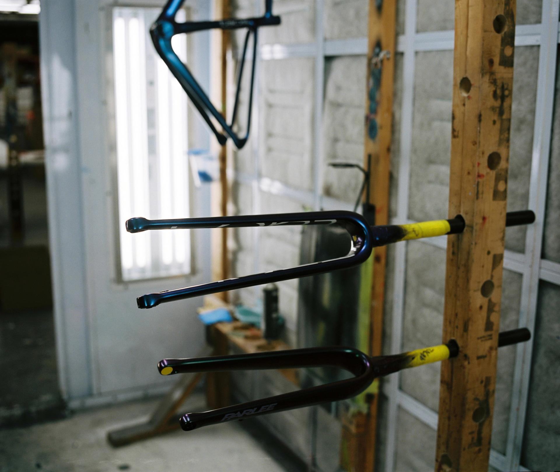 Three freshly painted bicycle forks suspended on a drying stand, their bright yellow tips contrasting against the neutral workshop interior.