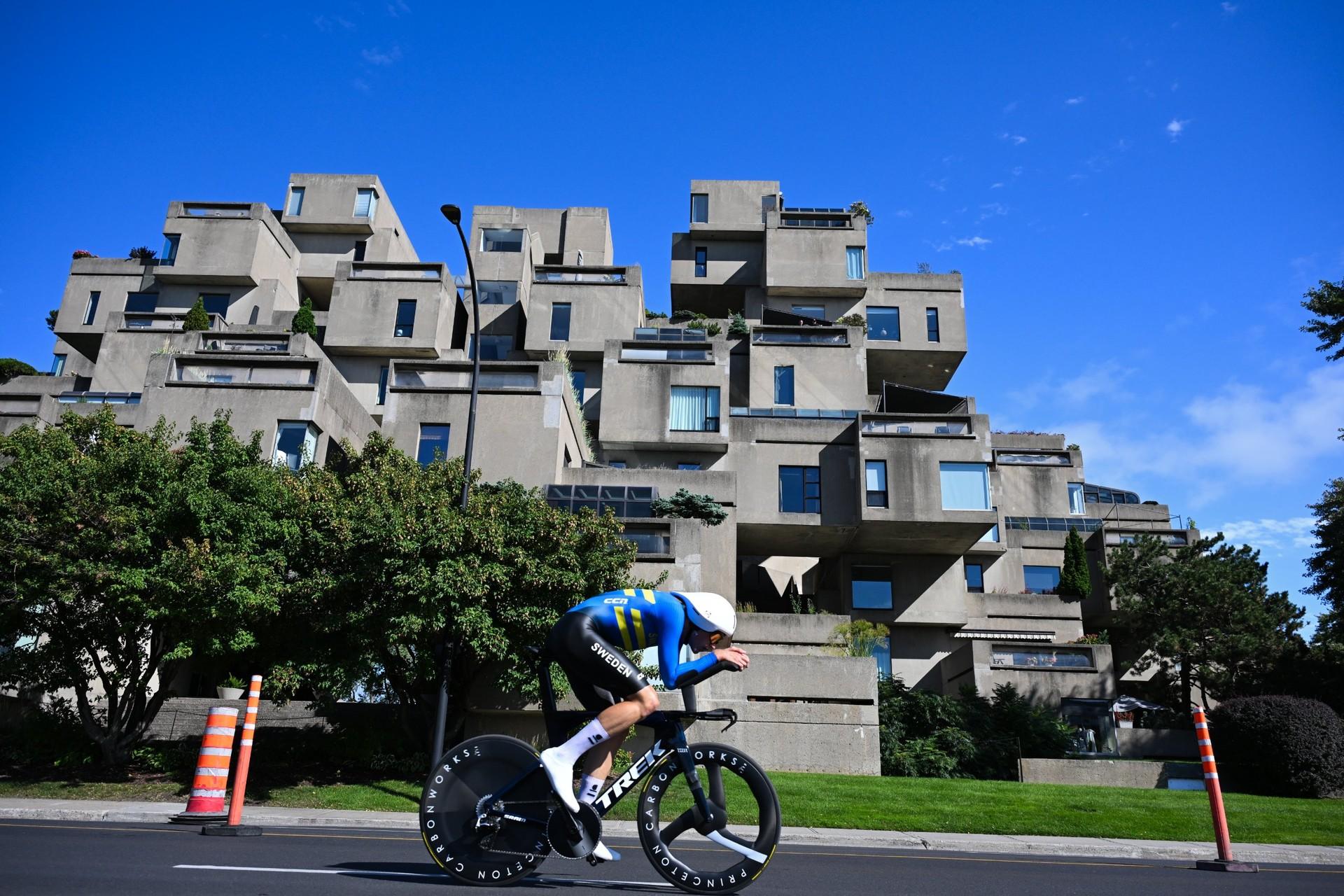 A cyclist in a blue kit rides past modern buildings and trees.