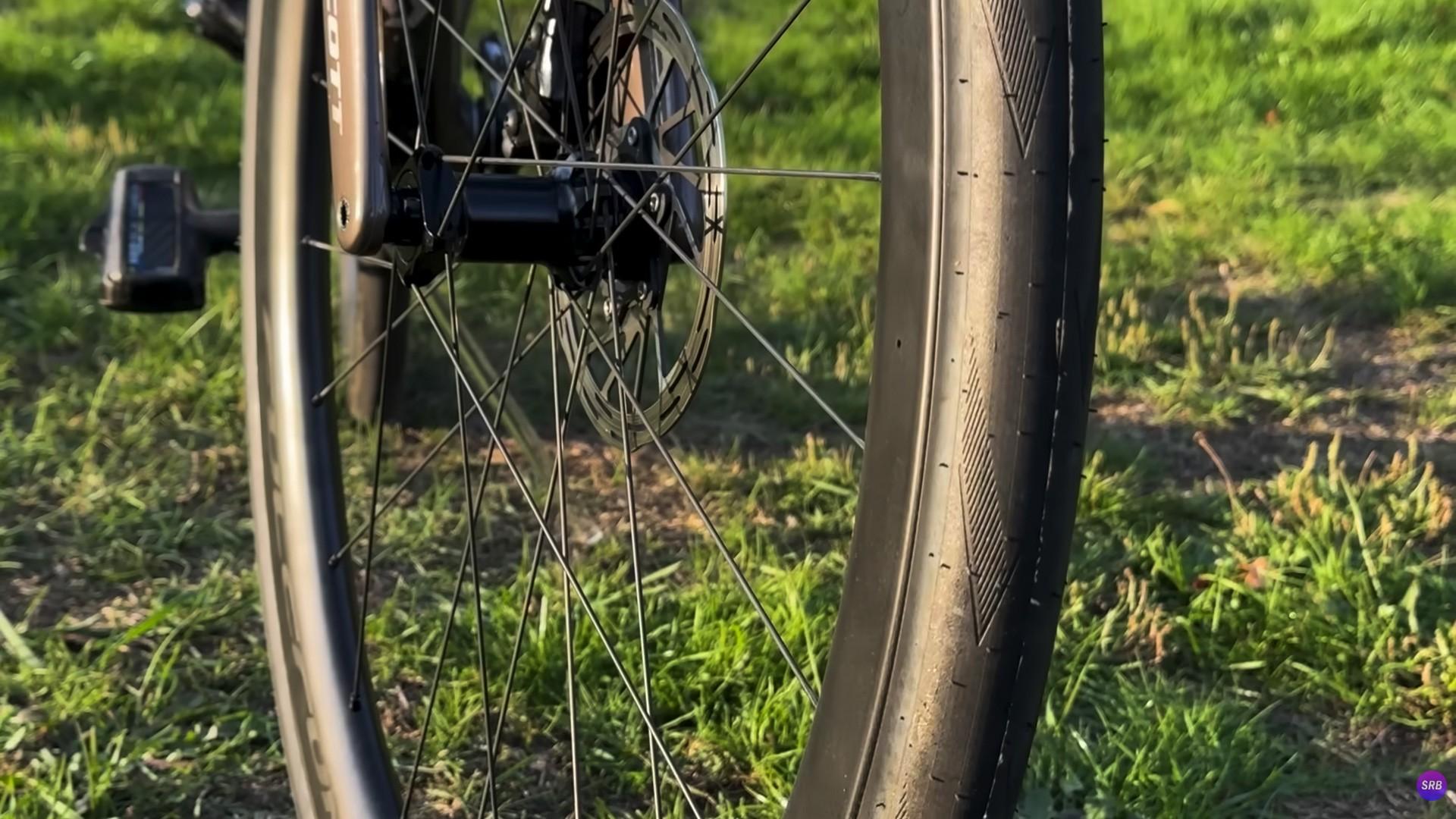 Close-up of a road bike front wheel featuring disc brakes and carbon rims on a grassy field at sunset.
