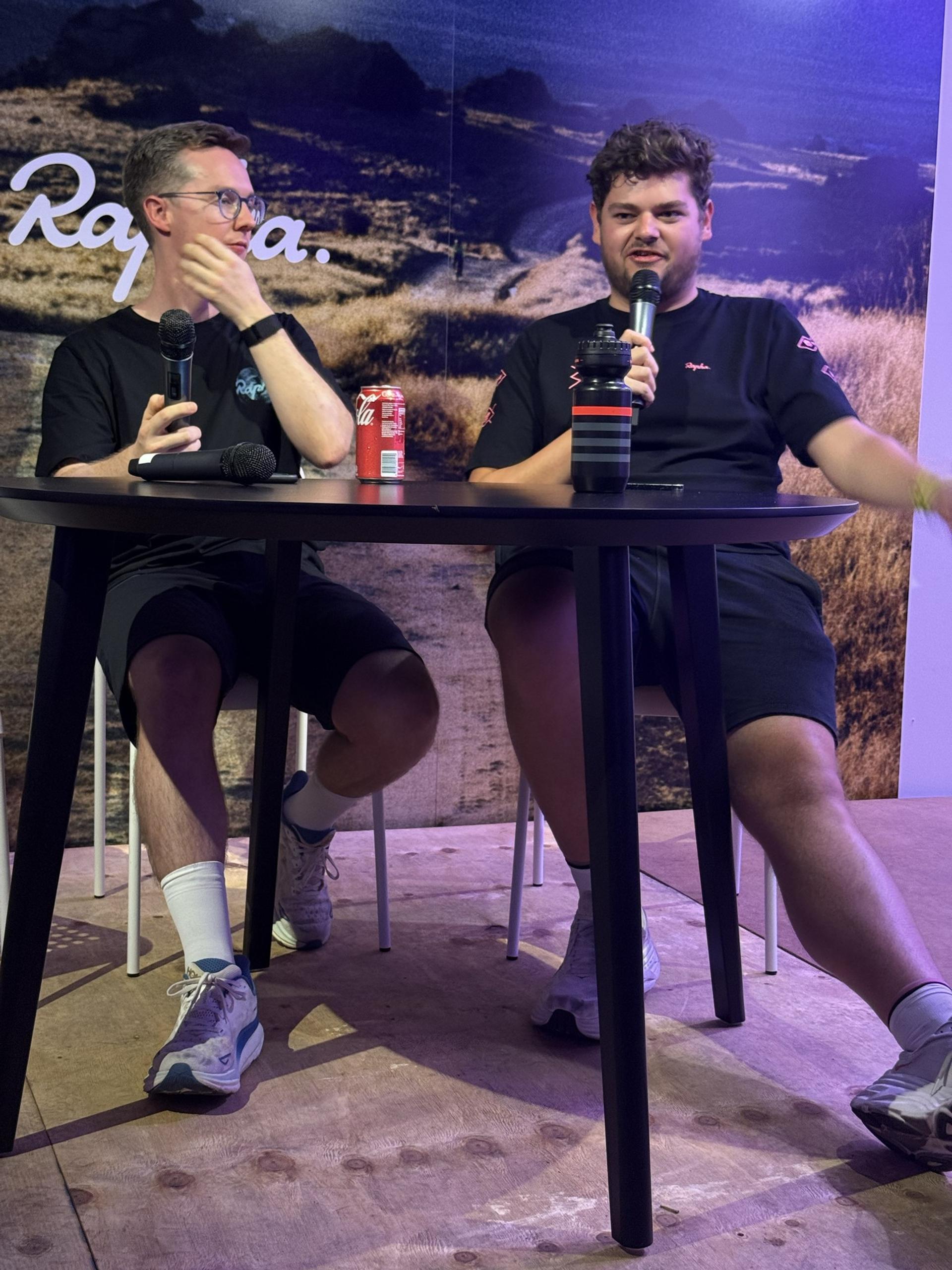 Two men seated at a panel, each speaking into microphones during a cycling event discussion