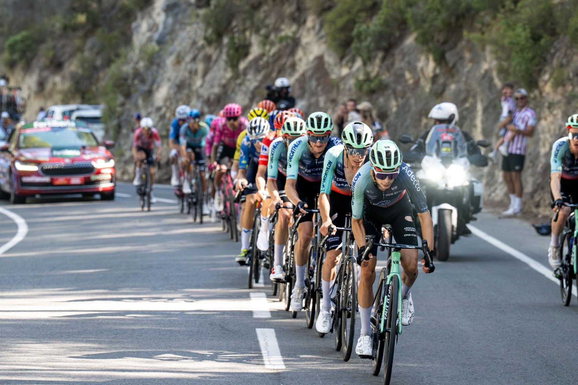 Cyclists climb a mountain road in a tight group, followed by team cars and a motorcycle.