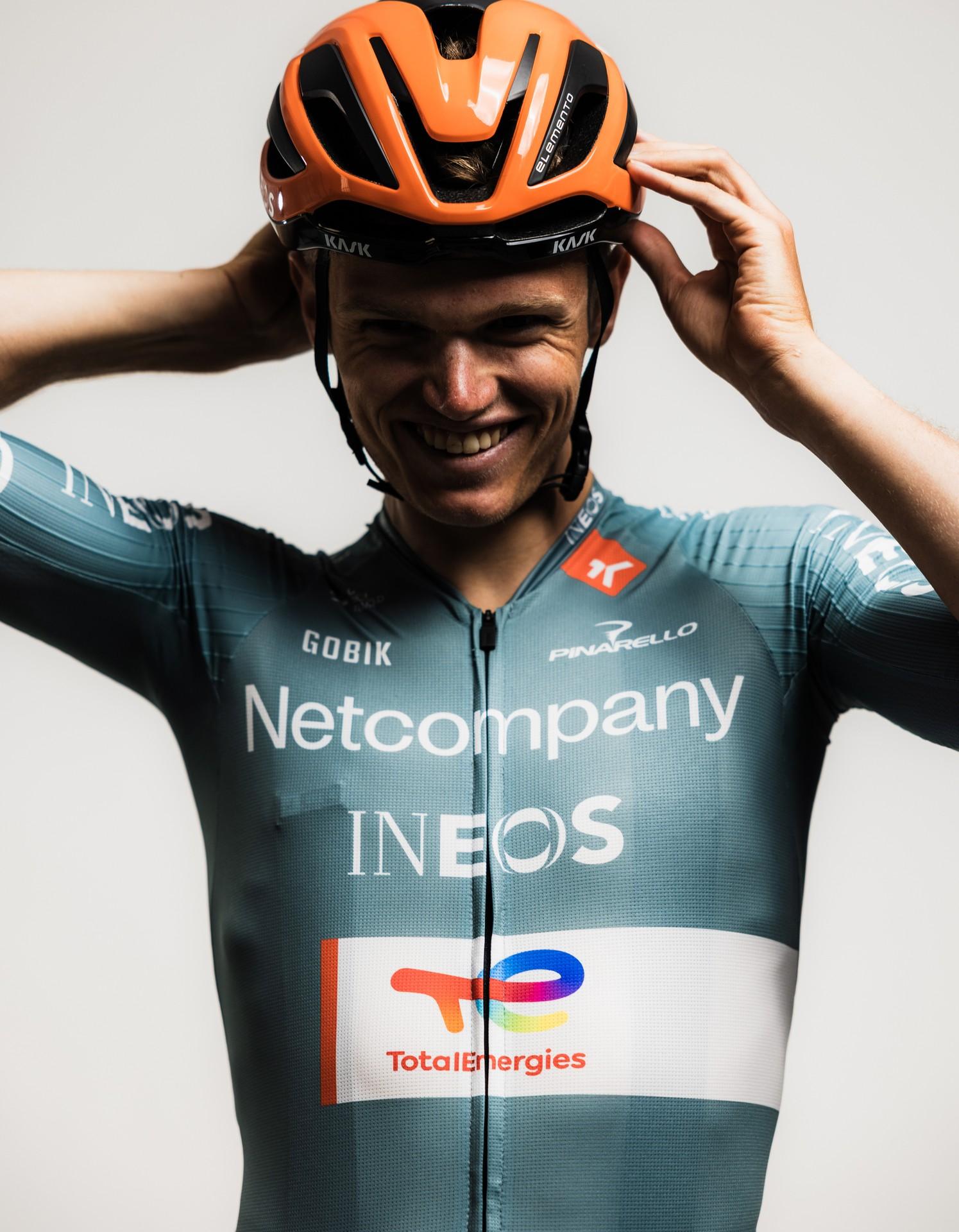 Cyclist in Team INEOS kit adjusts orange helmet and smiles during a celebratory moment