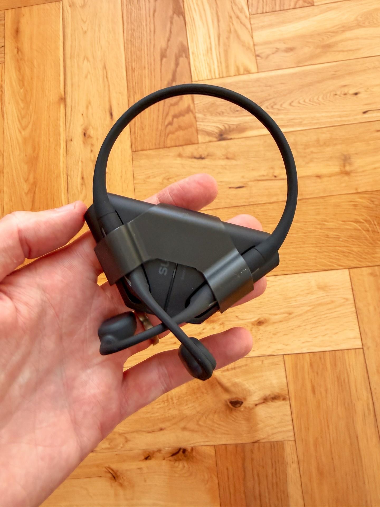 A hand holds the black Suunto Wing 2 bone-conduction headphones against a wooden floor.