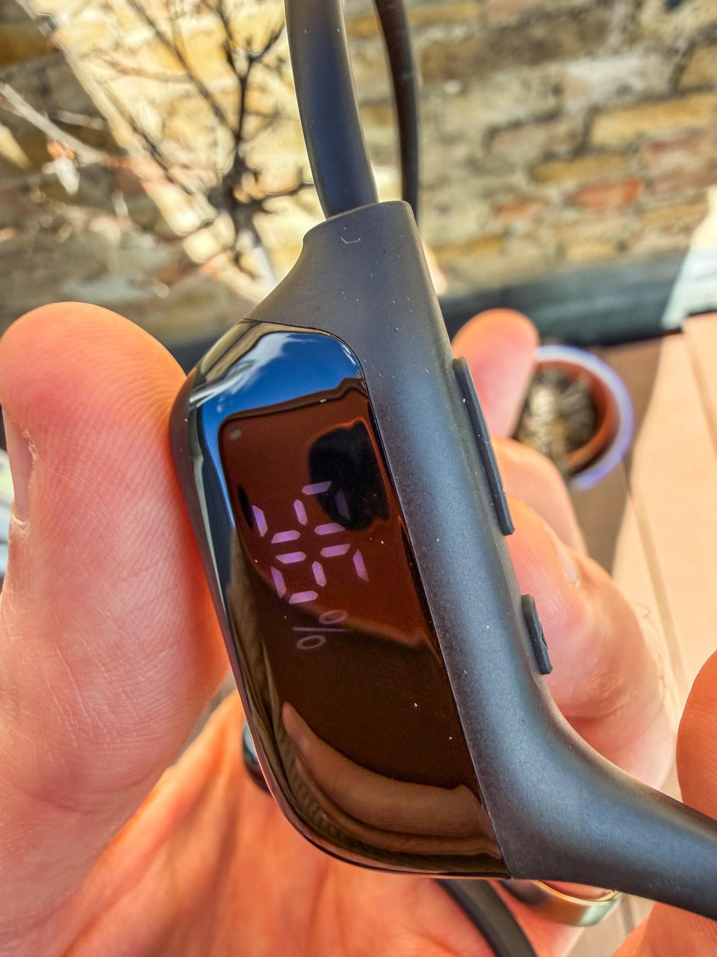 A hand holds the Zovimax bone-conduction headphones, showing their small status display and neckband.