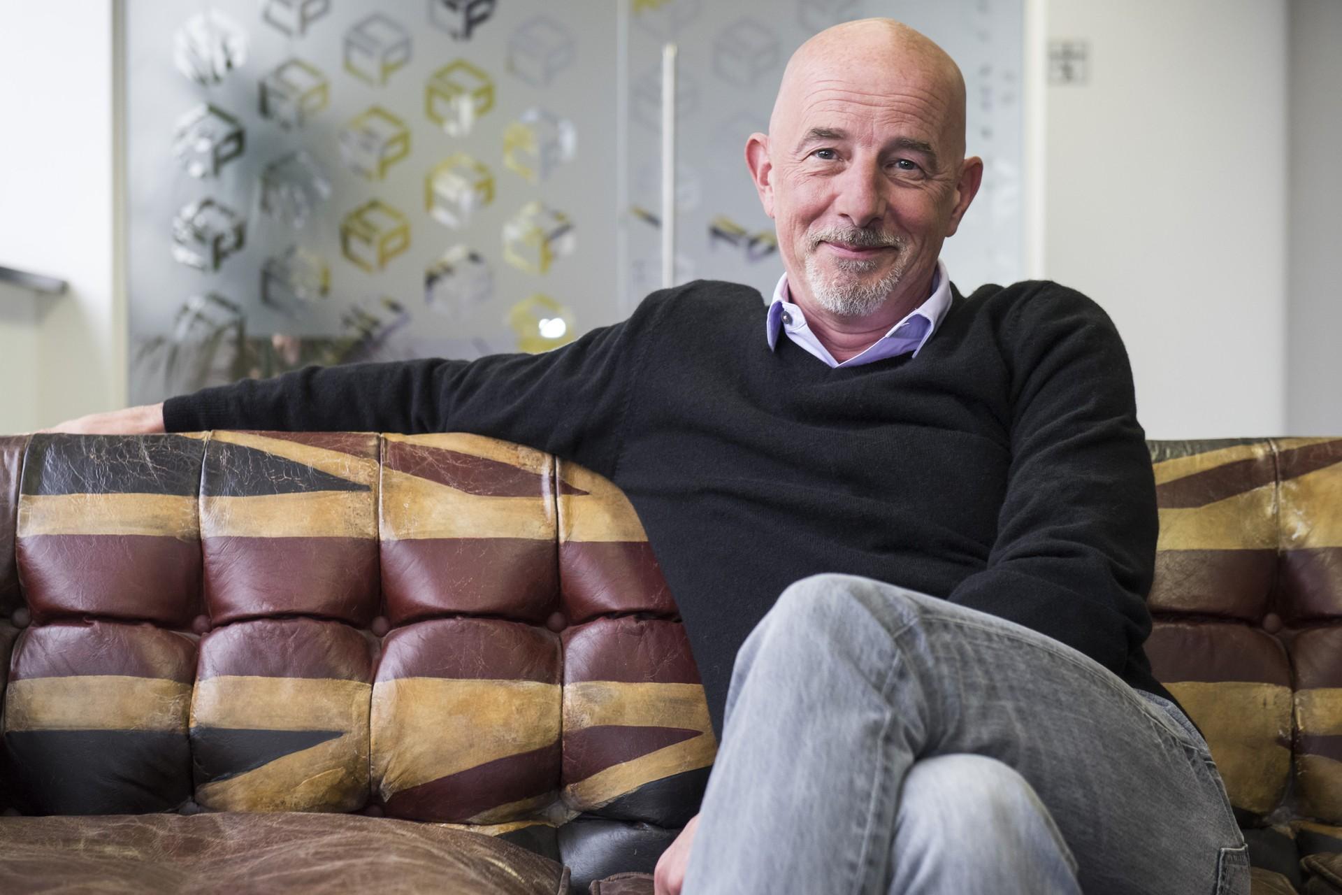 Ewan Kirk sits on a sofa with one leg crossed, wearing a black sweater and gray trousers.