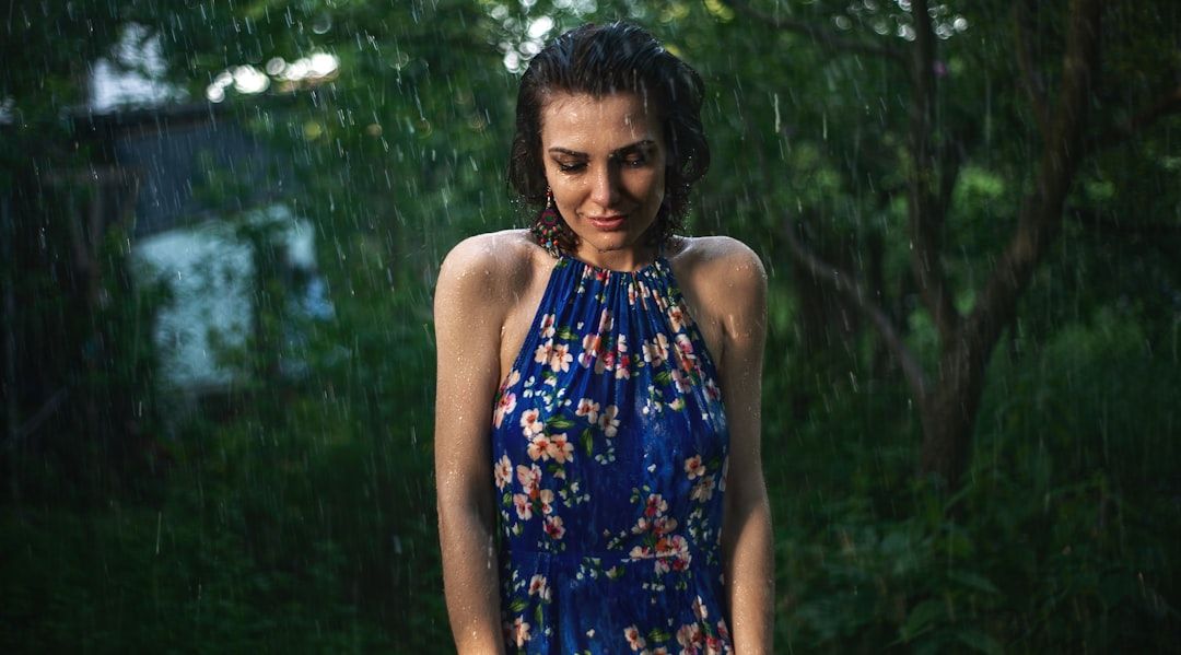 A young girl in a bright, patterned frock wearing a light raincoat and boots, smiling in a rainy outdoor setting.