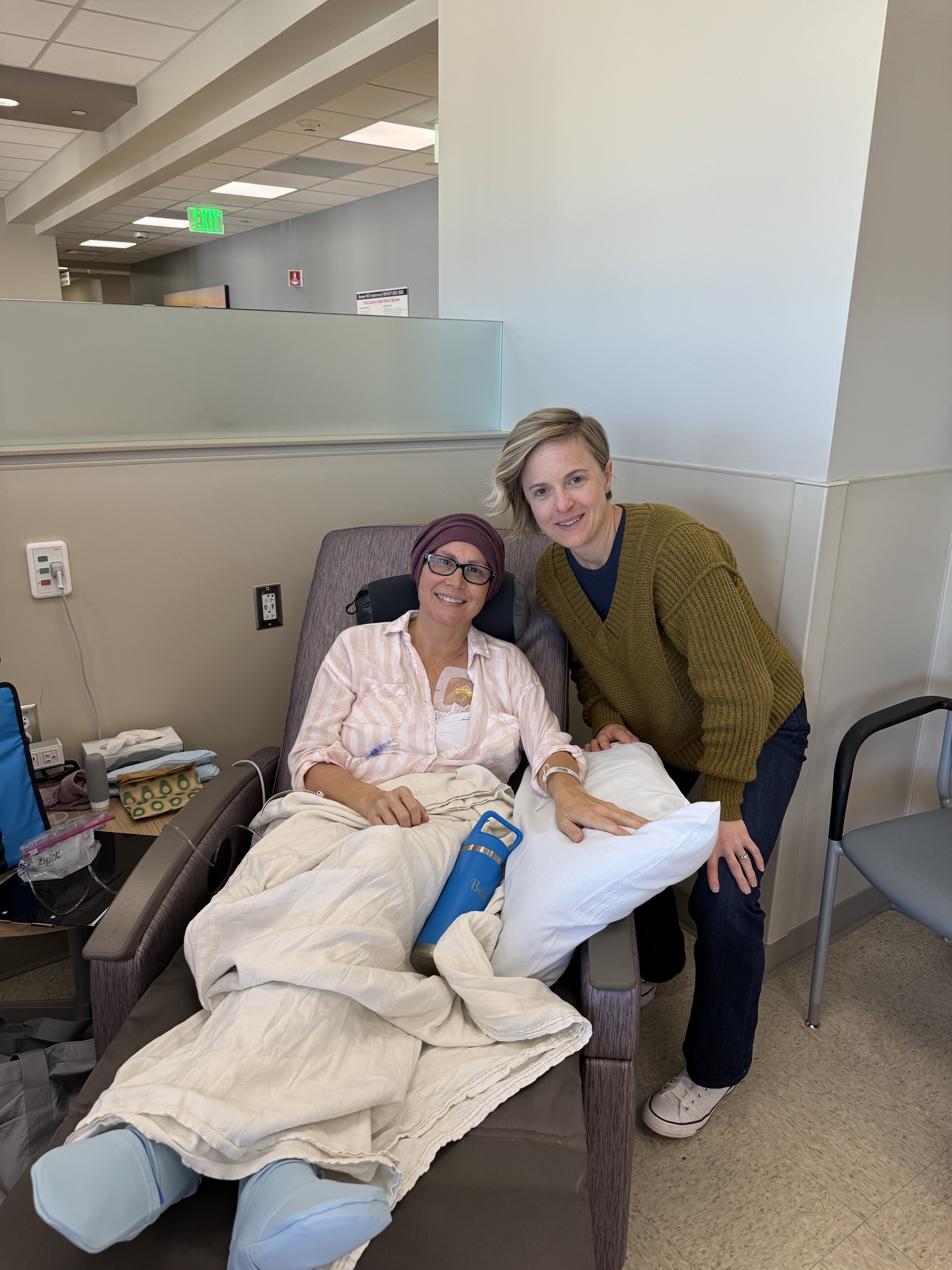 A woman seated in a medical recliner beside another woman in a clinic treatment room.